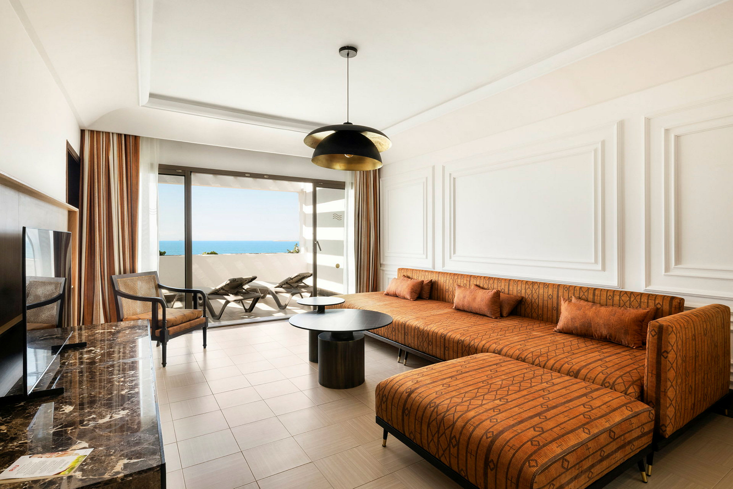 Et moderne hotellrom med en oransje sofa, et salongbord og utsikt mot havet fra en balkong. (AI-generert tekst)