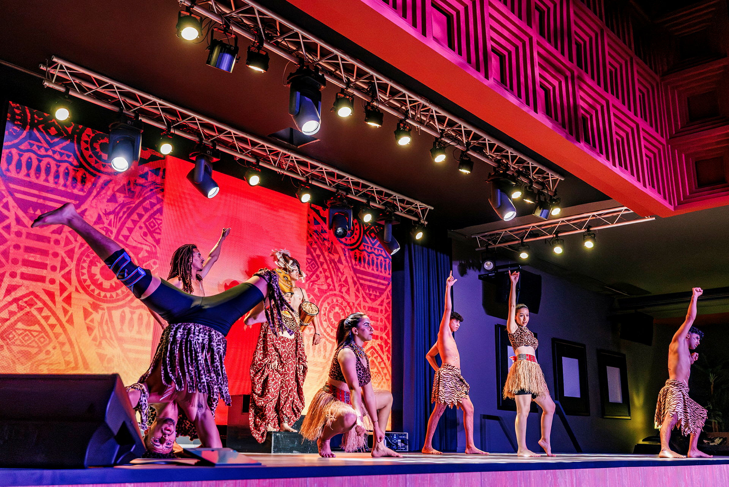 Gruppe av dansere på scenen med lys fargerik bakgrunn og tribal-inspirerte kostymer. (AI-generert tekst)