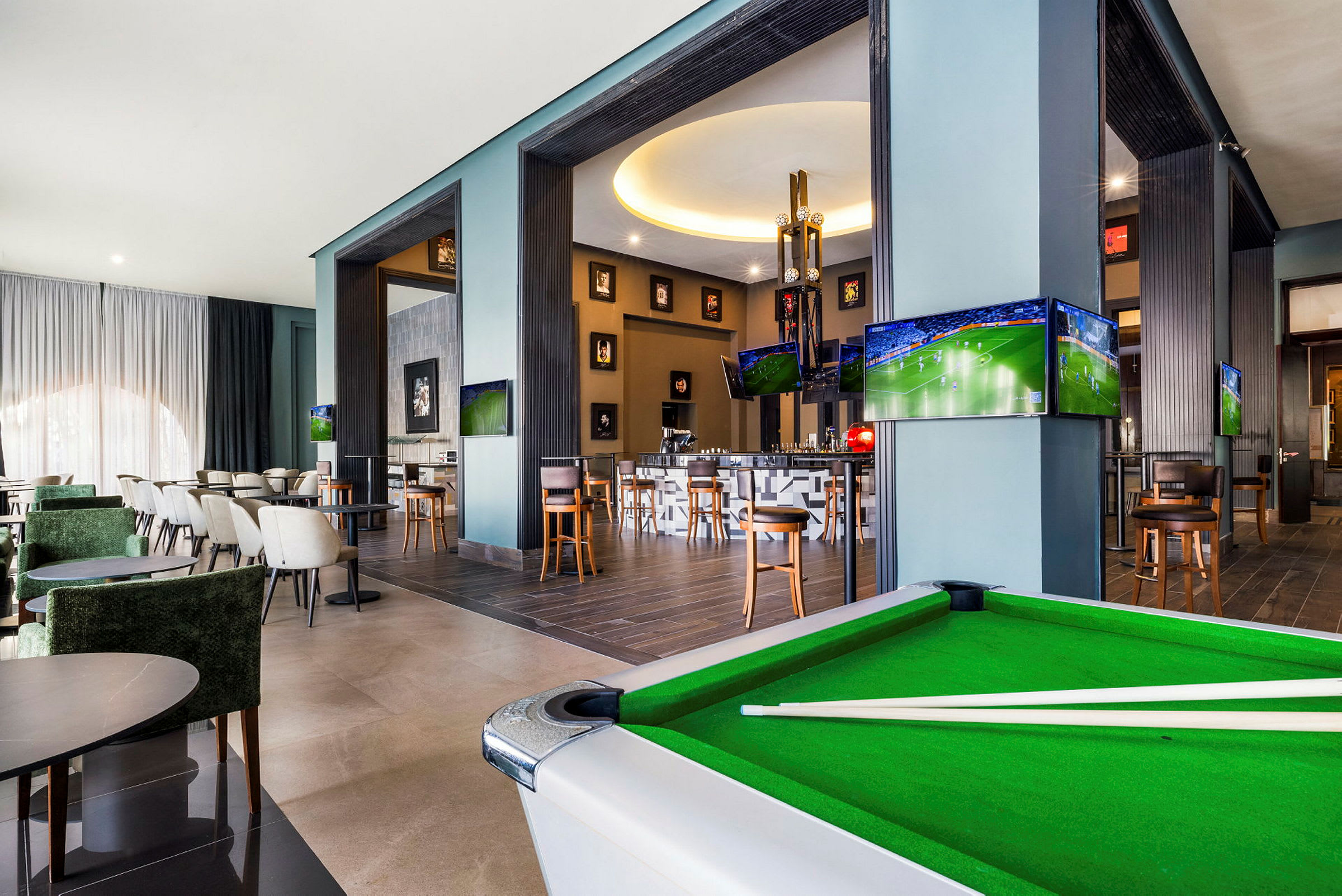 Interiør av en moderne lounge med sittegrupper, et biljardbord og flere TV-skjermer som viser sport. (AI-generert tekst)