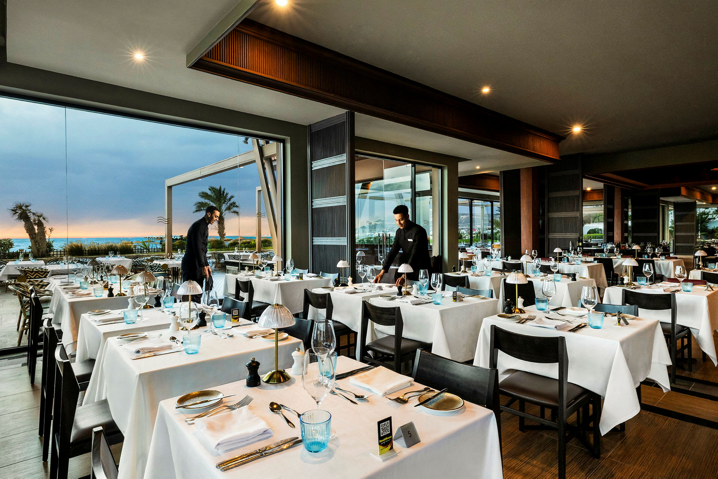 Interiørbilde av en restaurant med dekkede bord og store glassvinduer som viser utsikten mot havet og palmer. (AI-generert tekst)