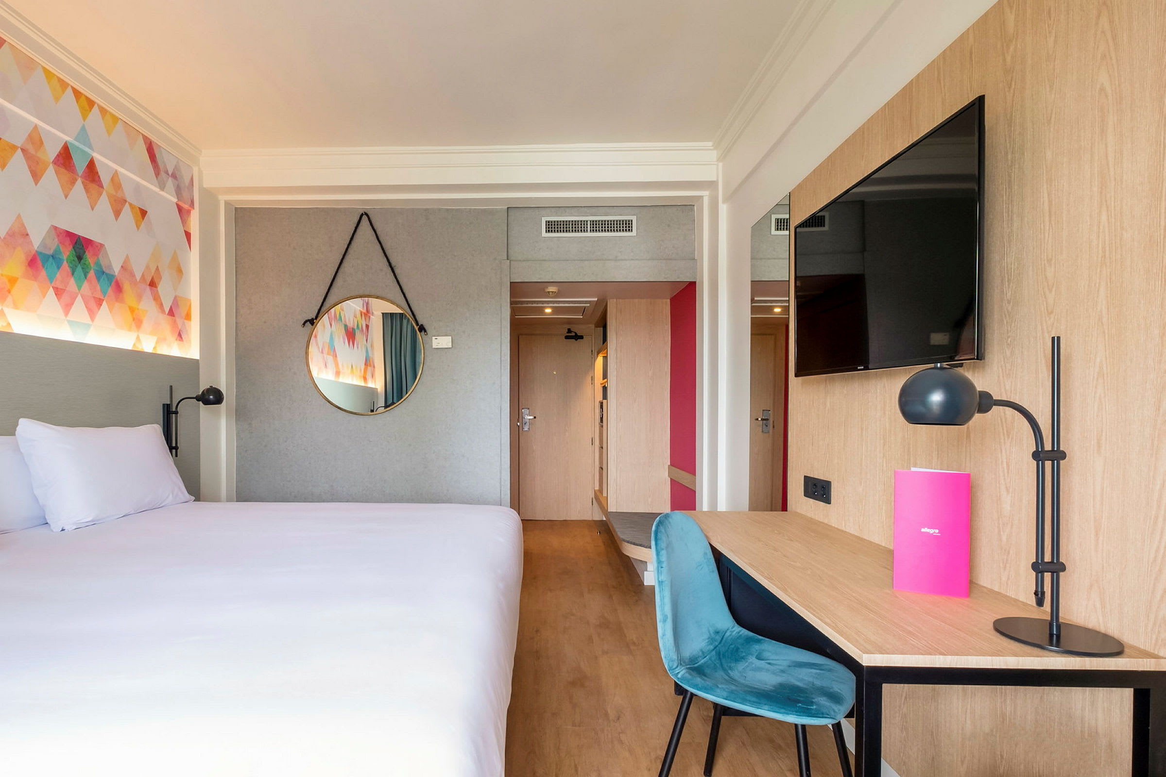 Et hotellrom med en seng, et skrivebord med stol og lampe, samt en veggmontert TV. (AI-generert tekst)