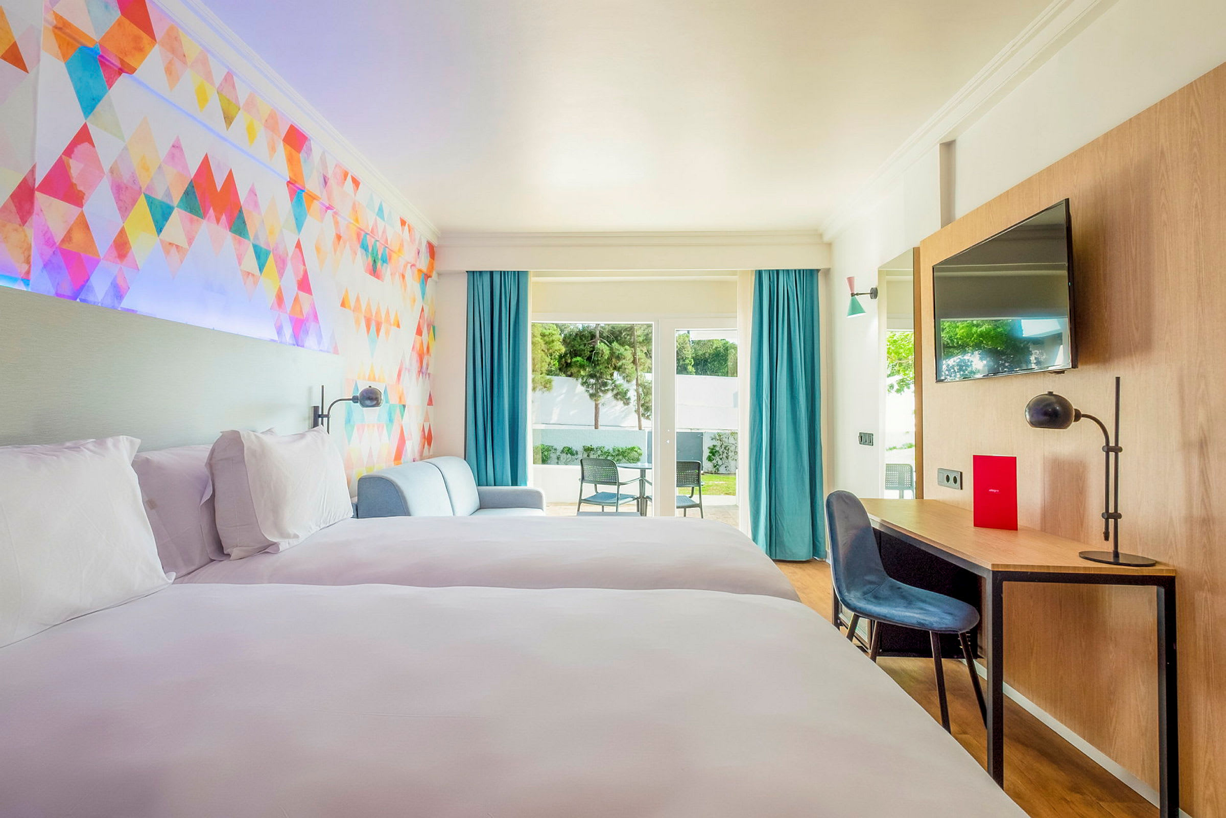 Et hotellrom med to senger, en fargerik veggdekorasjon, sittegruppe, skrivebord og tilgang til en terrasse med stoler. (AI-generert tekst)