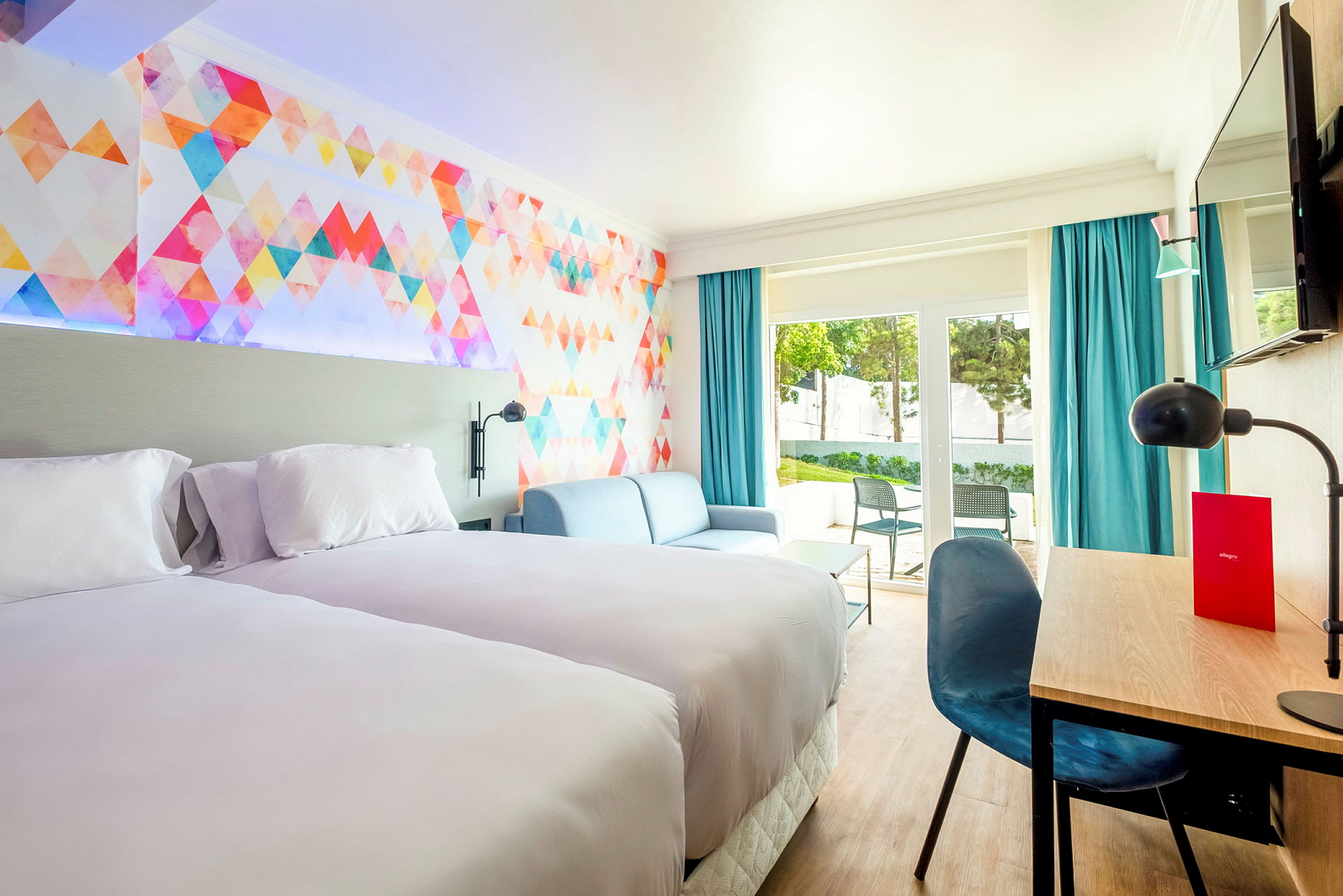 Hotellrom med to senger, sofa og skrivebord. Fargerikt veggdesign og store vinduer. (AI-generert tekst)