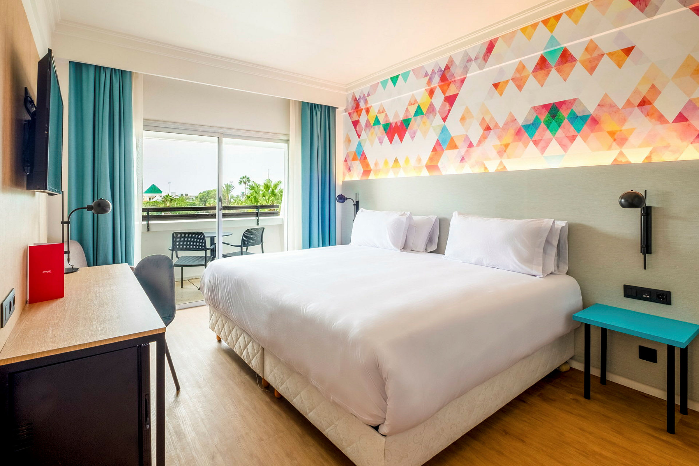 Et hotellrom med en dobbeltseng, fargerikt veggdesign, skrivebord, og balkong med to stoler. (AI-generert tekst)