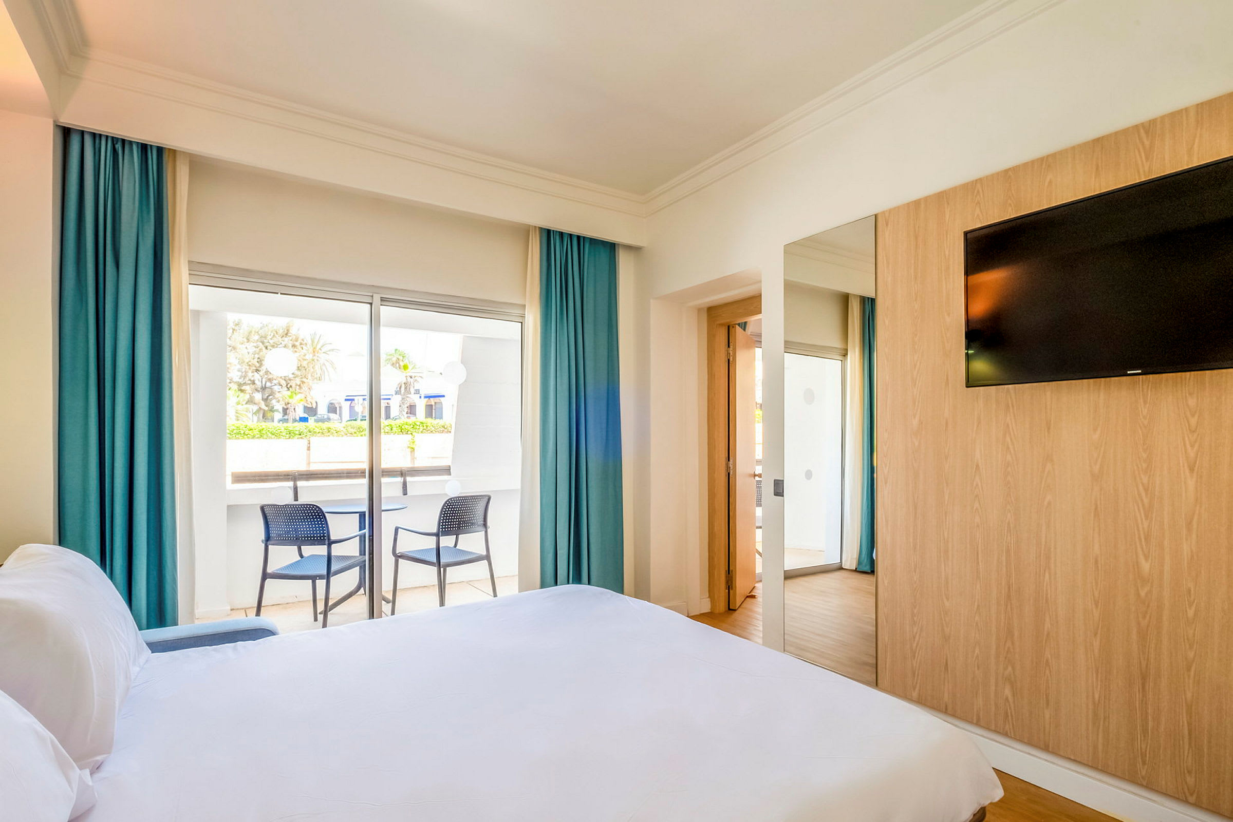 Et hotellrom med en seng, en veggmontert TV og en balkong med to stoler. (AI-generert tekst)