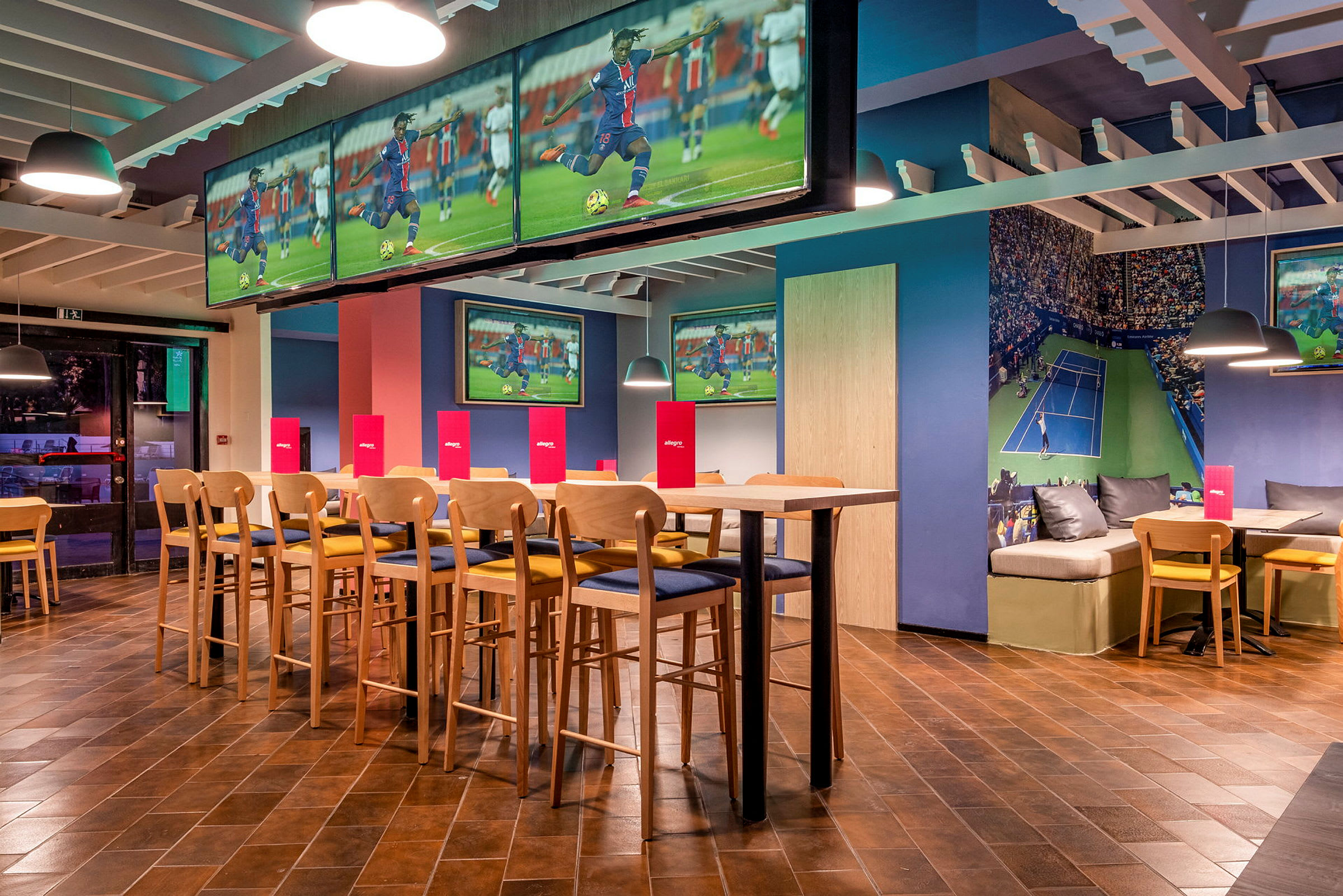 Interiør av en sportsbar med TV-skjermer som viser fotball og tennis, sitteplasser og bord. (AI-generert tekst)