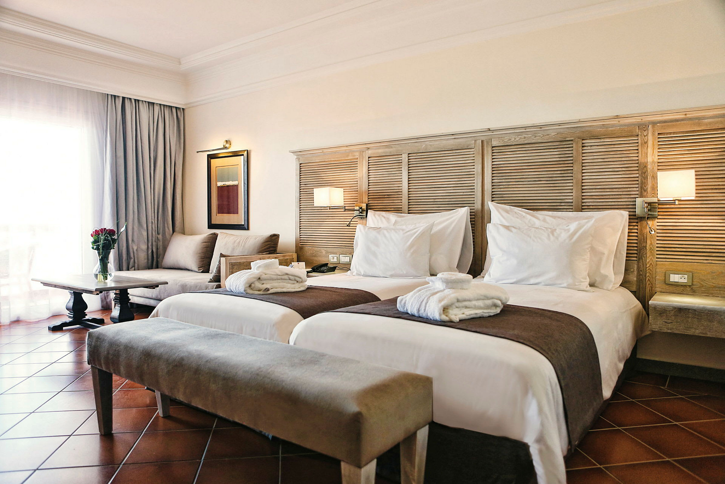 Et hotellrom med to separate senger, en sofa, et bord med en blomster vase og et vindu med gardiner. (AI-generert tekst)