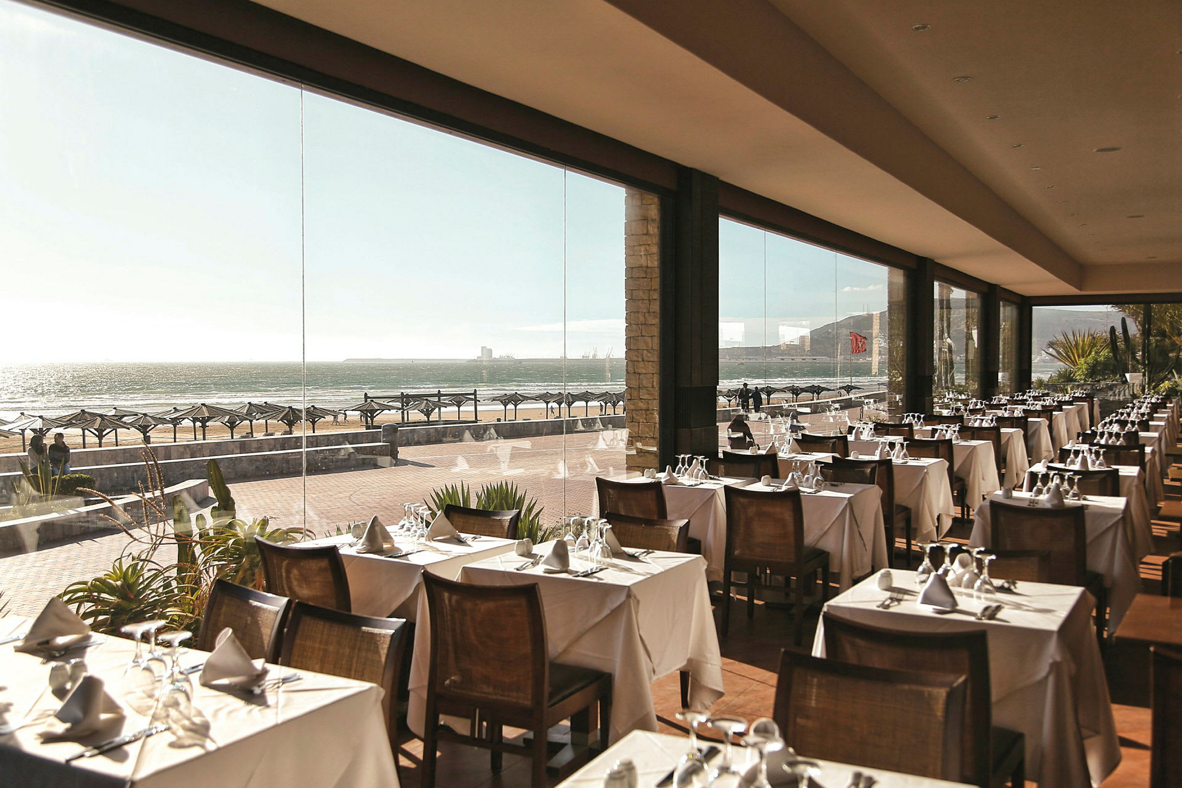 Restaurant med dekket bord og glassvegger med utsikt over stranden. (AI-generert tekst)