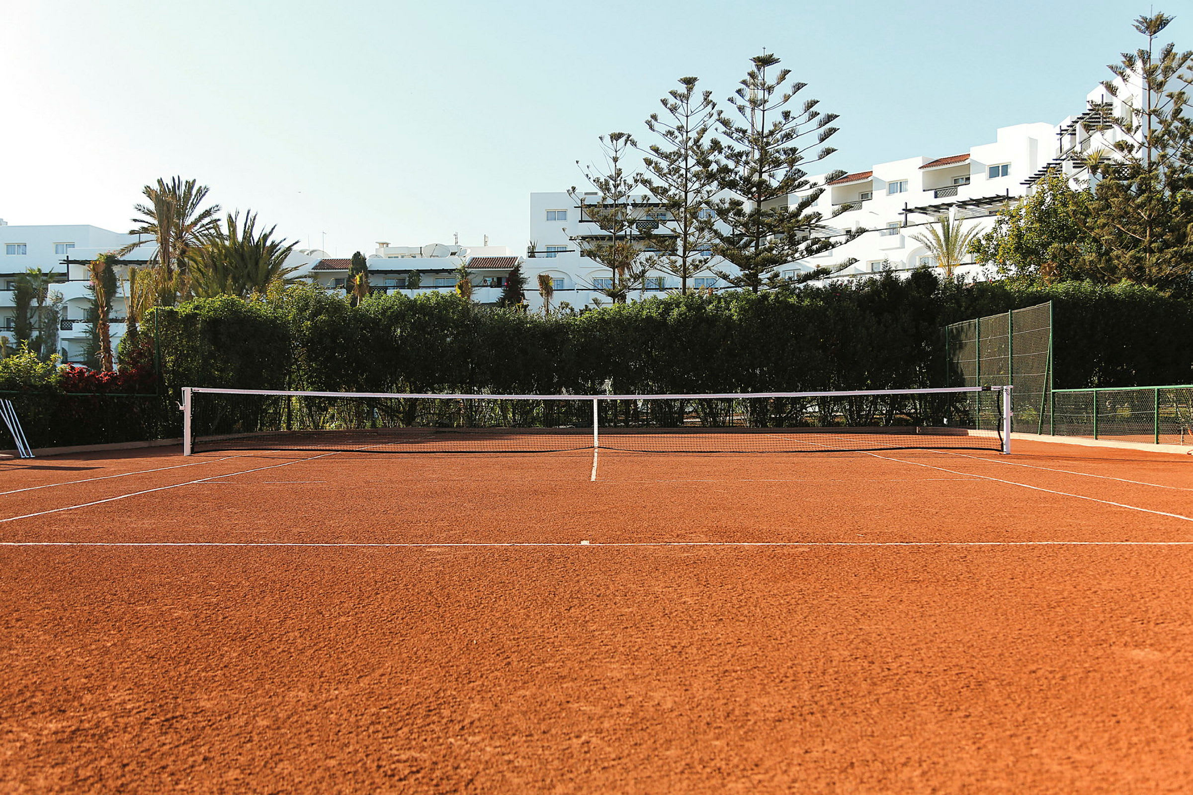 Bilde av en tennisbane med grusunderlag, omgitt av trær og bygninger i bakgrunnen. (AI-generert tekst)