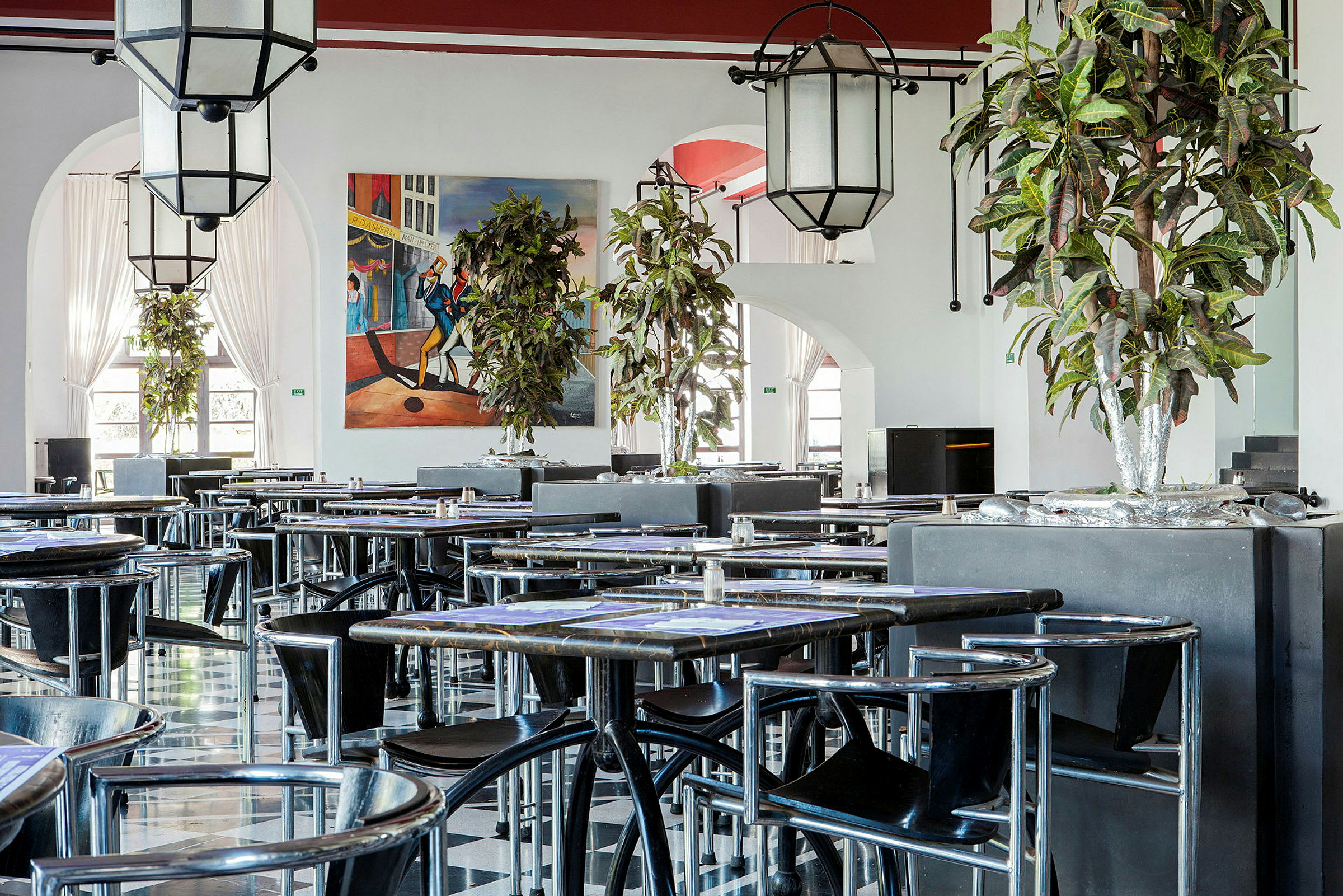 En innendørs restaurant med bord og stoler. Detaljer som planter og en fargerik veggmaleri er synlige. (AI-generert tekst)