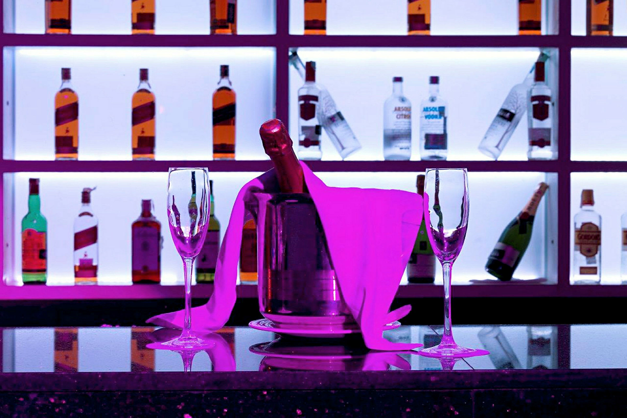 Et bord med to tomme champagneglass og en champagneflaske i en ishink, omgitt av rosa belysning. I bakgrunnen er det hyller med flasker. (AI-generert tekst)