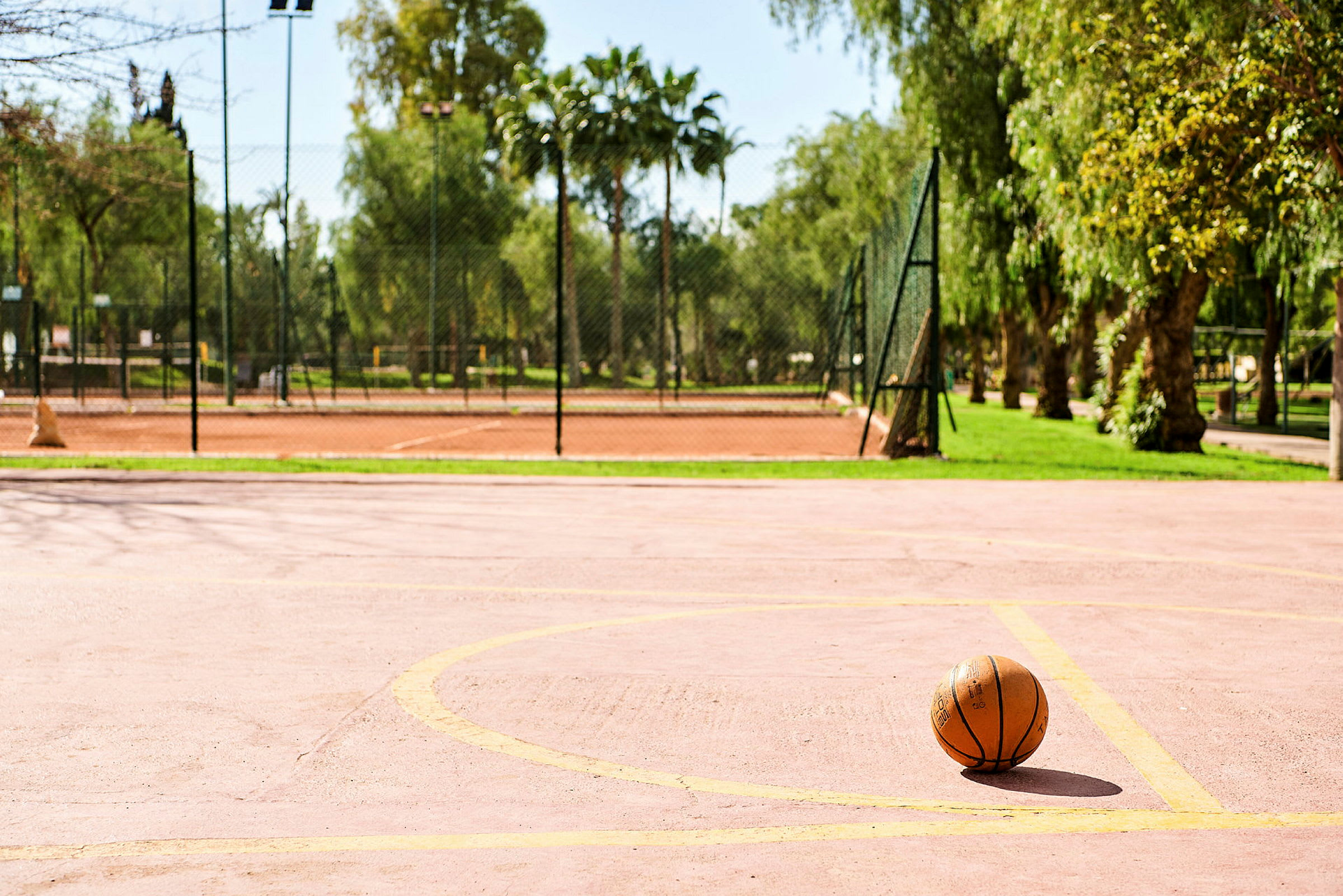 En basketball på en tom basketballbane med trær og gjerde i bakgrunnen. (AI-generert tekst)