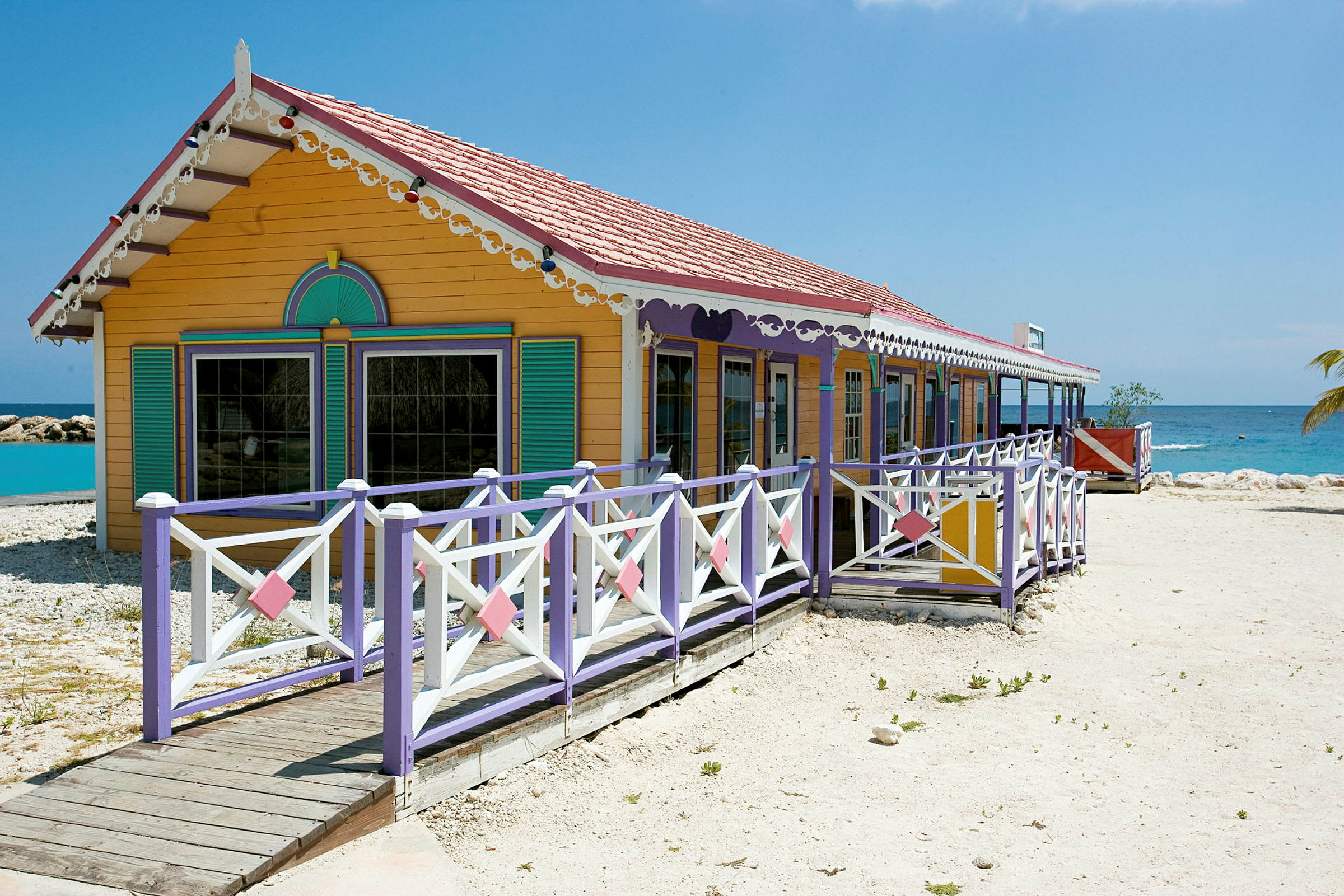 Et fargerikt hus med dekorasjoner og veranda, plassert ved en strand med klart blått vann. (AI-generert tekst)