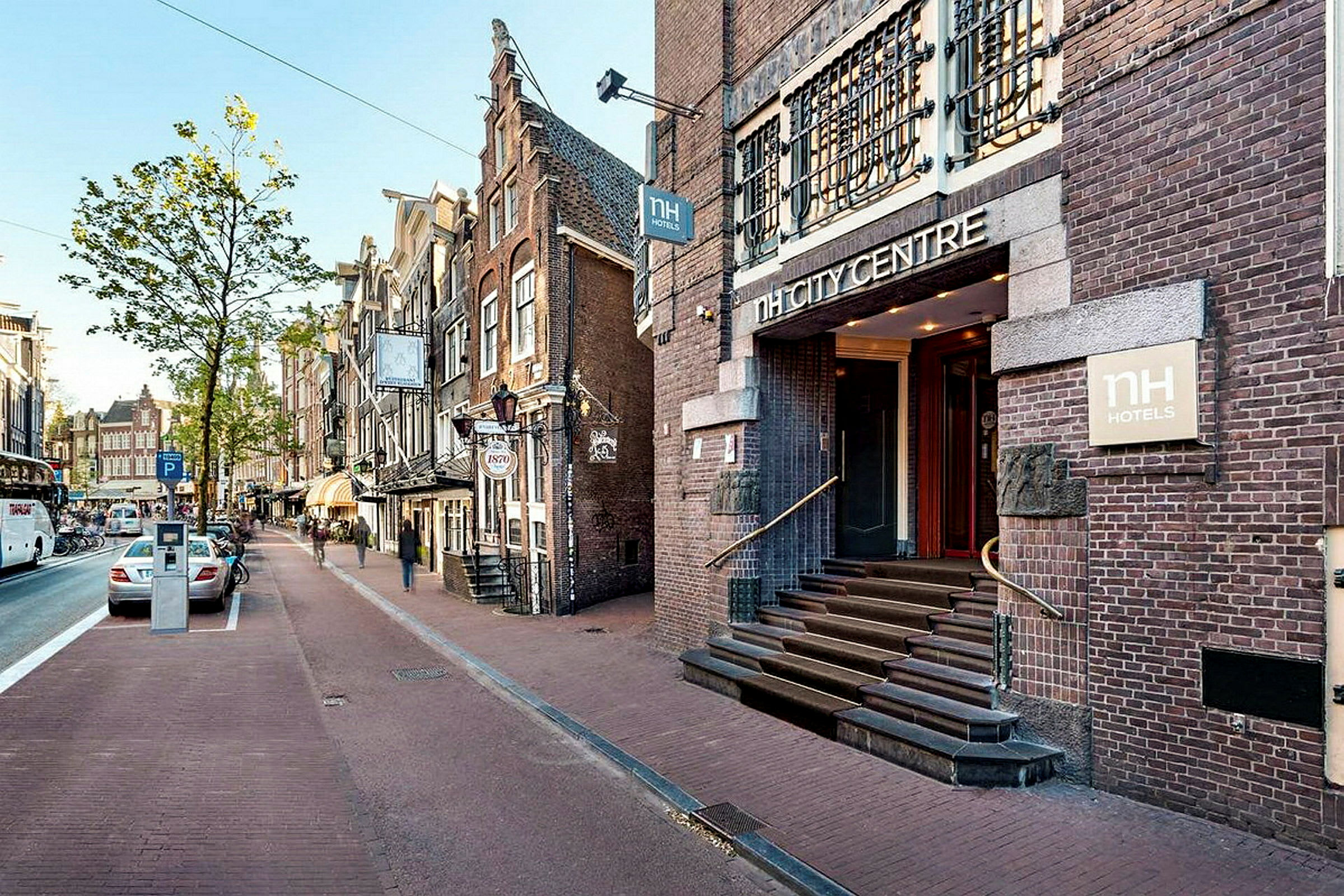 En gate med parkerte biler og gangvei, omkranset av bygninger i tradisjonell arkitektur hvor hotellet NH City Centre har en synlig inngang. (AI-generert tekst)