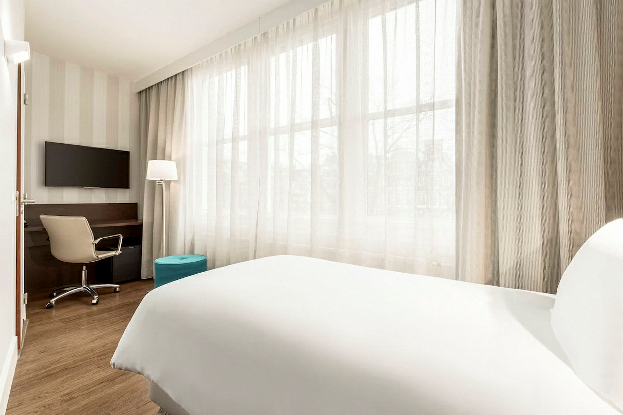 Et lyst hotellrom med en seng, skrivebord og stol samt en TV. Store vinduer dekket med lyse gardiner. (AI-generert tekst)