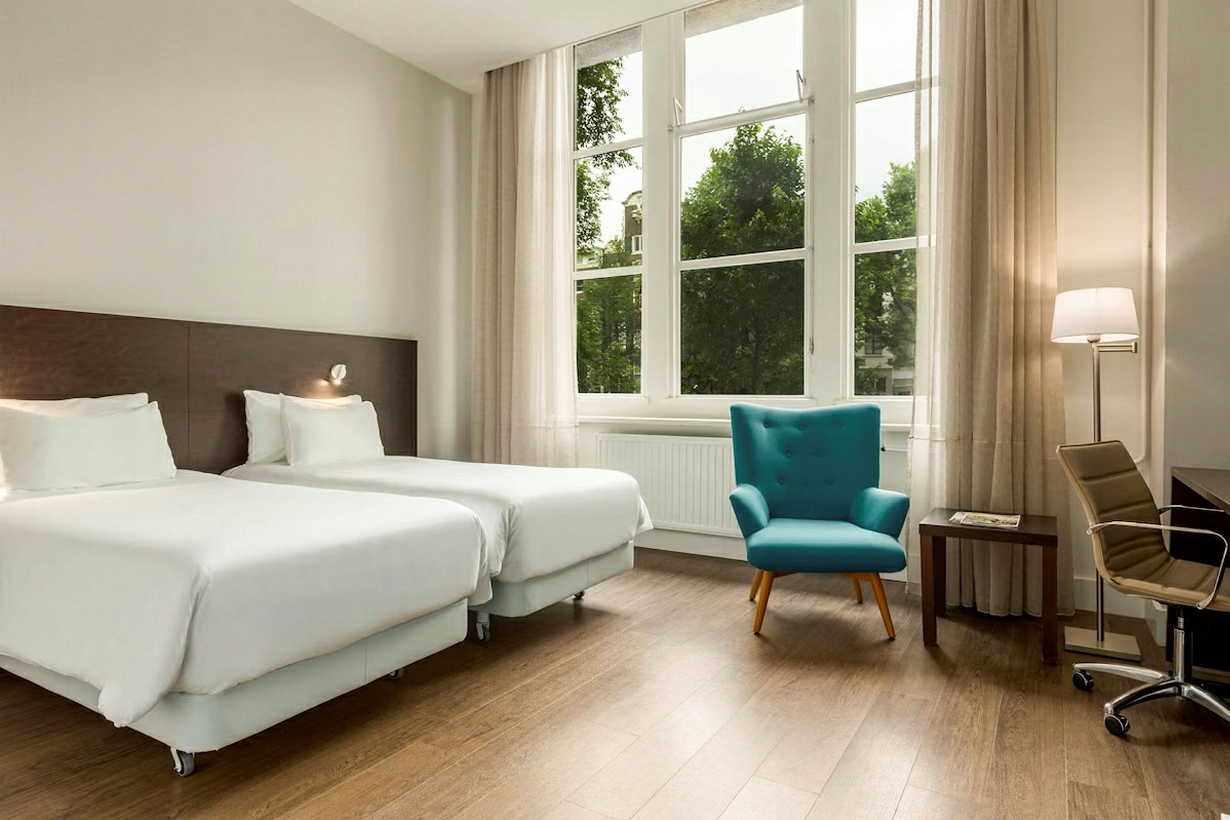 Et hotellrom med to separate senger, en turkis lenestol og et skrivebord med stol. Vinduet har gardiner og utsikt mot det grønne. (AI-generert tekst)