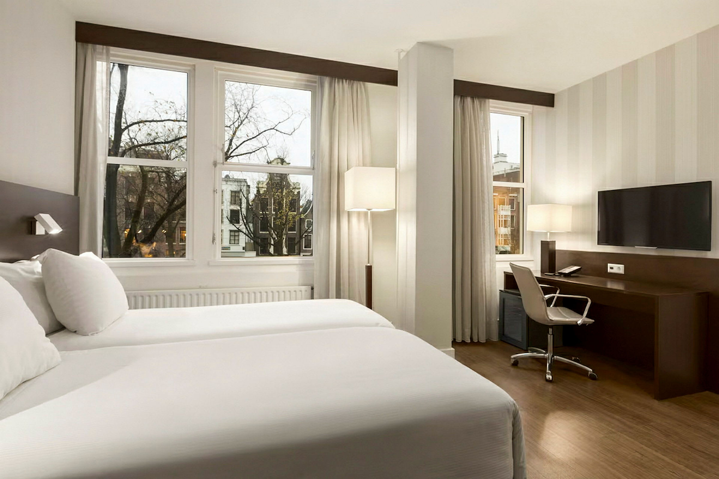 Hotellrom med to enkeltsenger, skrivebord, stol og TV, samt store vinduer med utsikt mot en bymiljø. (AI-generert tekst)