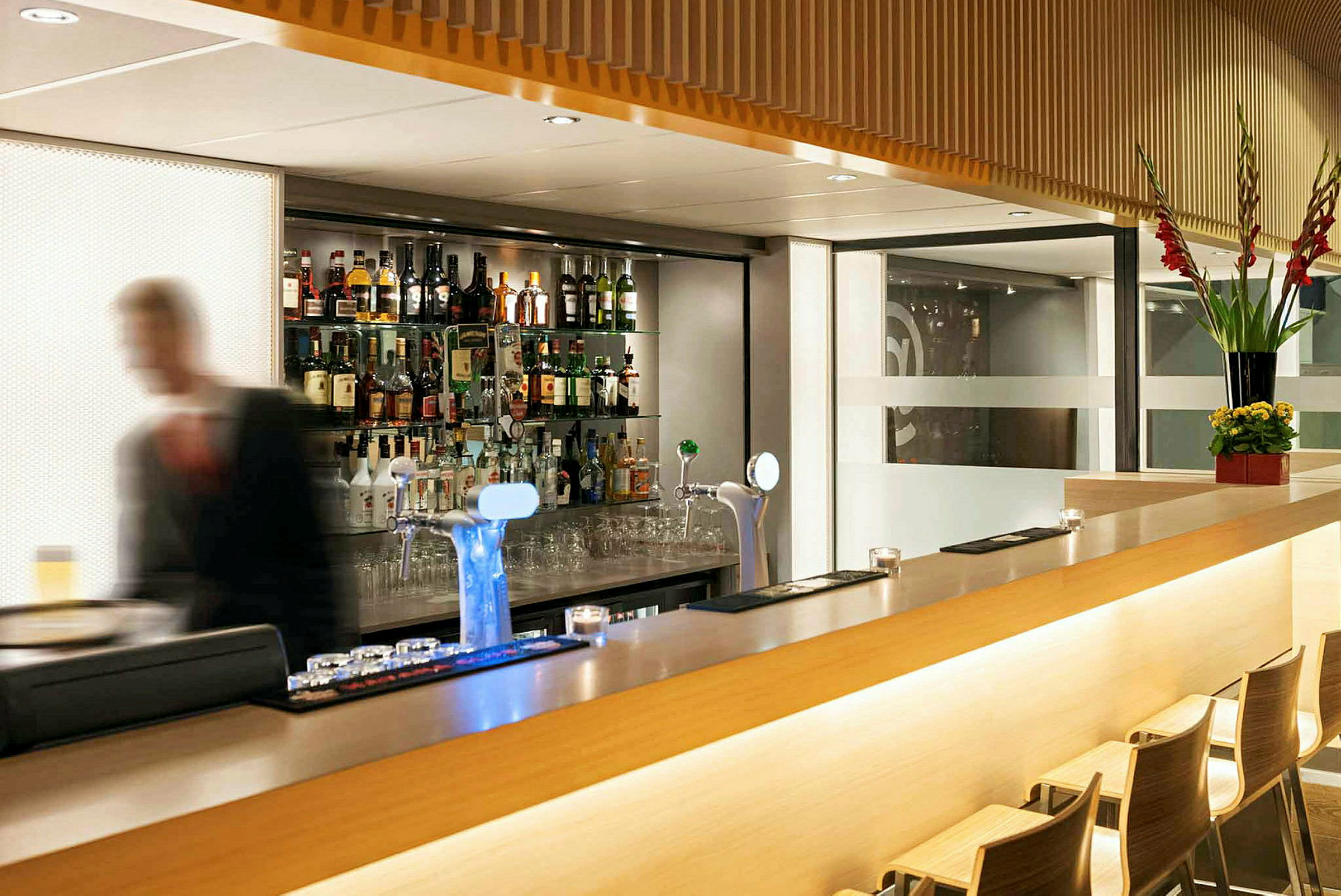 En moderne bar med et utvalg flasker på hyller bak samt en uskarp person i forgrunnen. (AI-generert tekst)