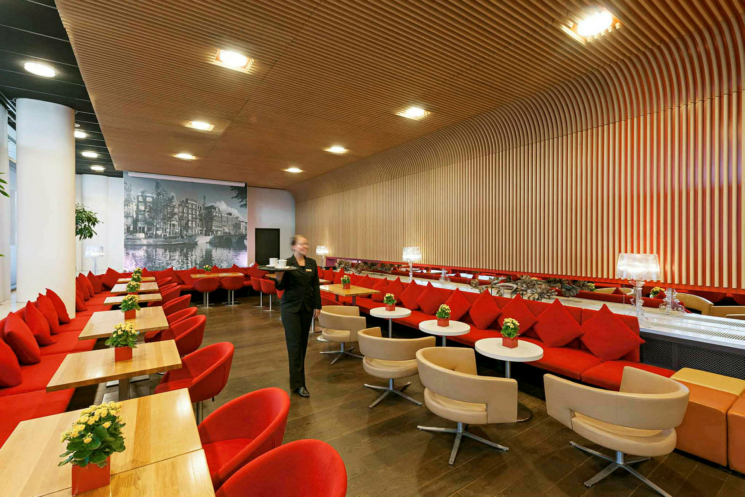 En moderne restaurant med røde og beige sitteplasser, en stor vegg med trepaneler og et bilde av byen i bakgrunnen. Bord og stoler er plassert i rader. (AI-generert tekst)