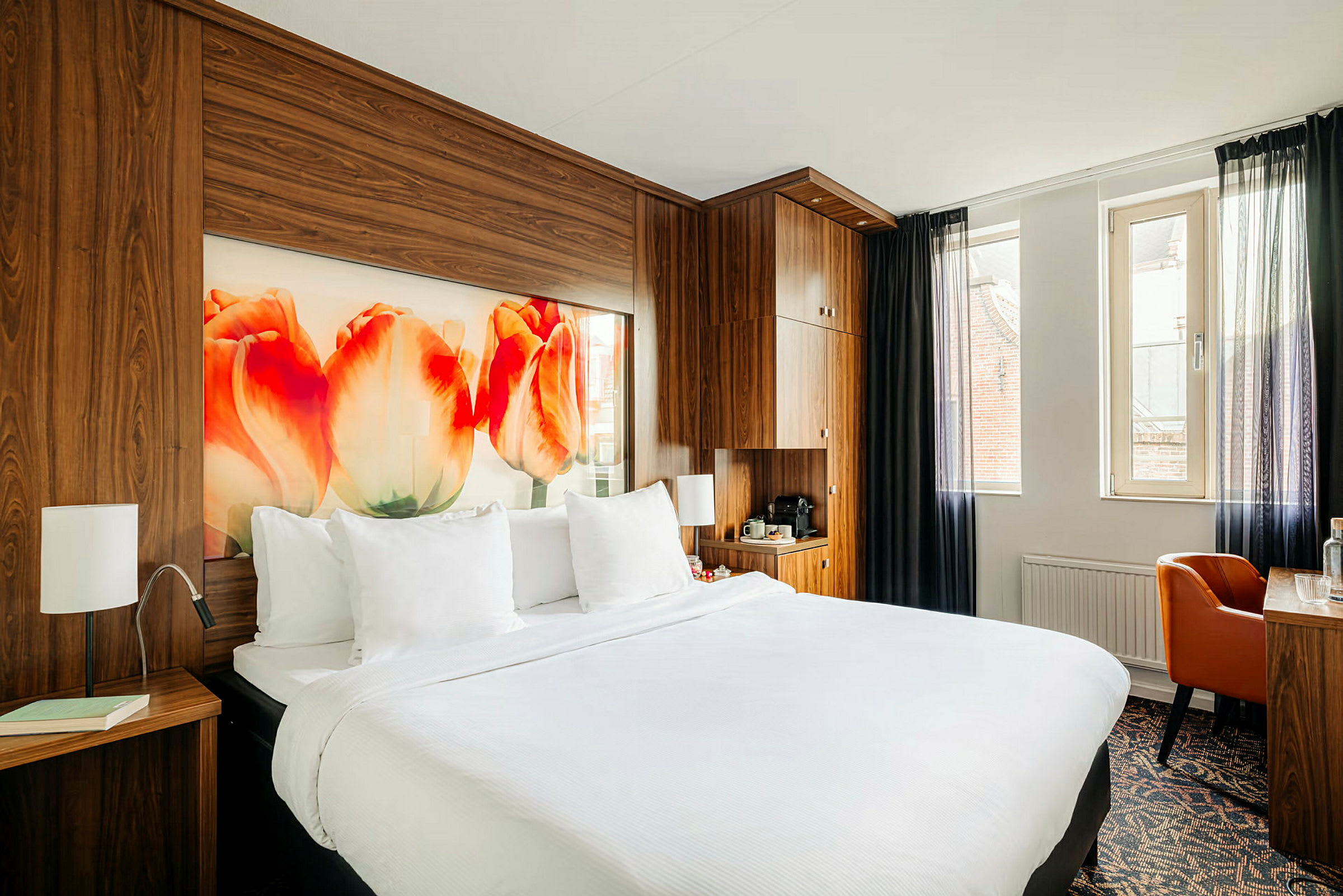 Hotellrom med en dobbeltseng, trepaneler, tulipanmotiv på veggen og et skrivebord ved vinduet. (AI-generert tekst)