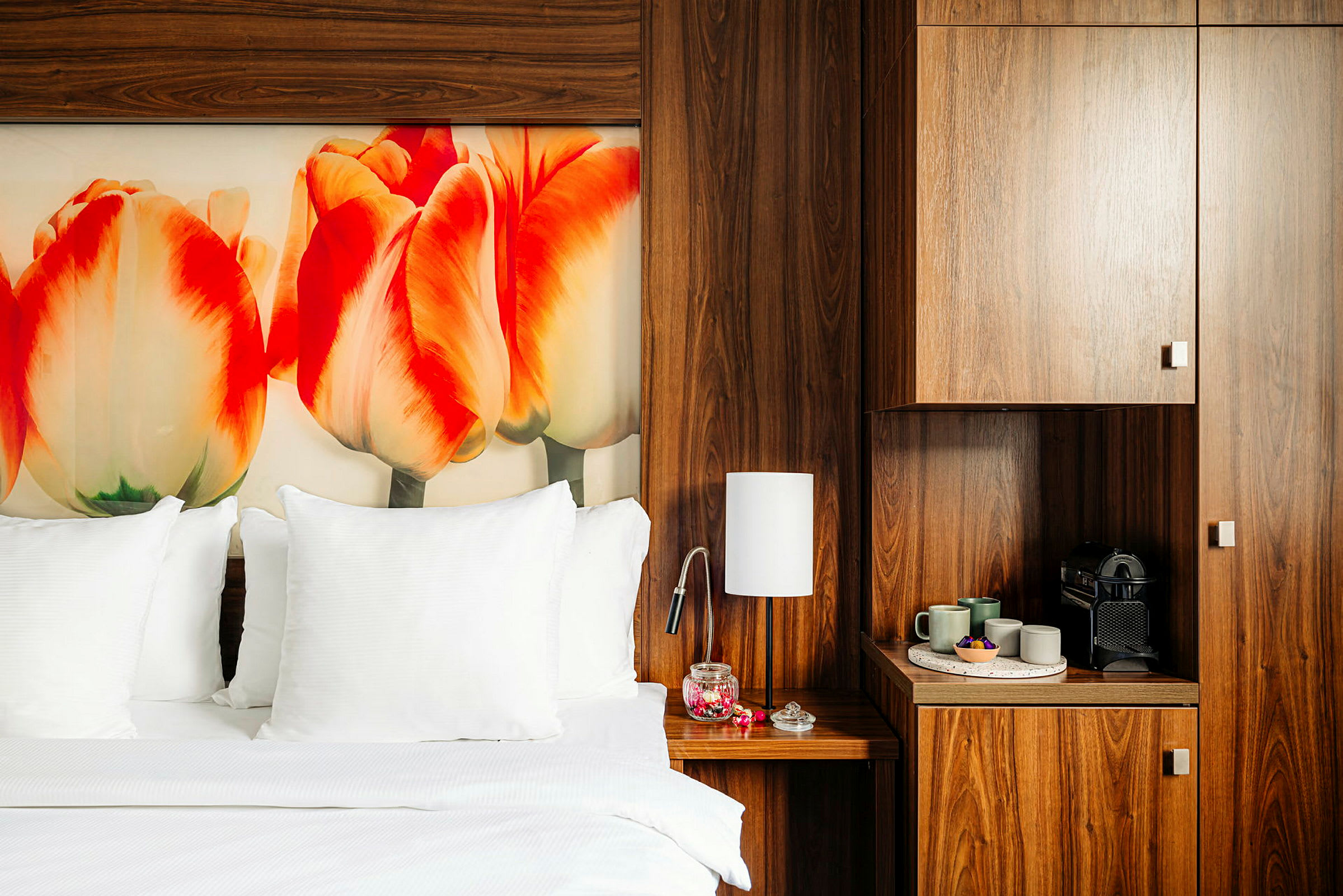 Et hotellrom med en stor seng med hvitt sengetøy og puter. På veggen er det et fargerikt bilde av tulipaner. Rommet har mørke tremøbler, inkludert et nattbord med en lampe og en kaffestasjon. (AI-generert tekst)