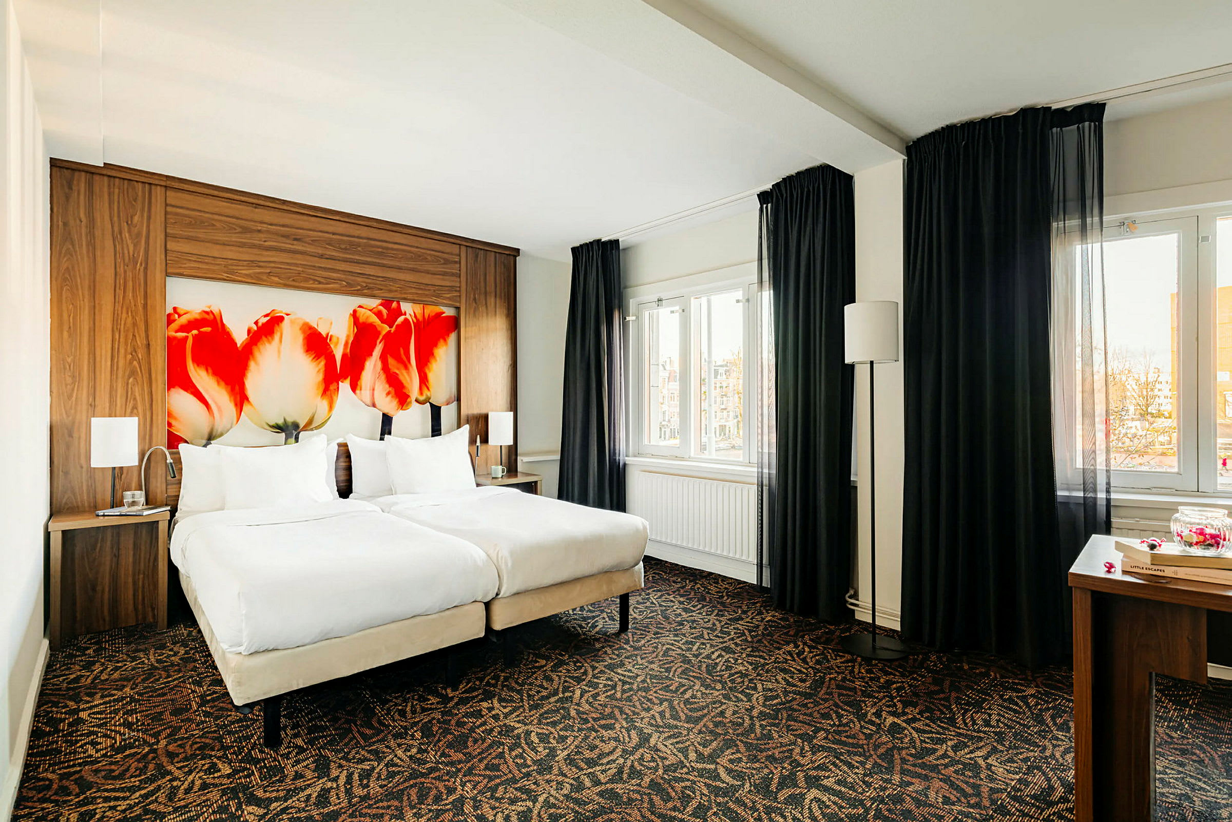 Et hotellrom med to enkeltsenger og en sengegavl med tulipanmotiv. Det finnes mørke gardiner ved vinduene. (AI-generert tekst)
