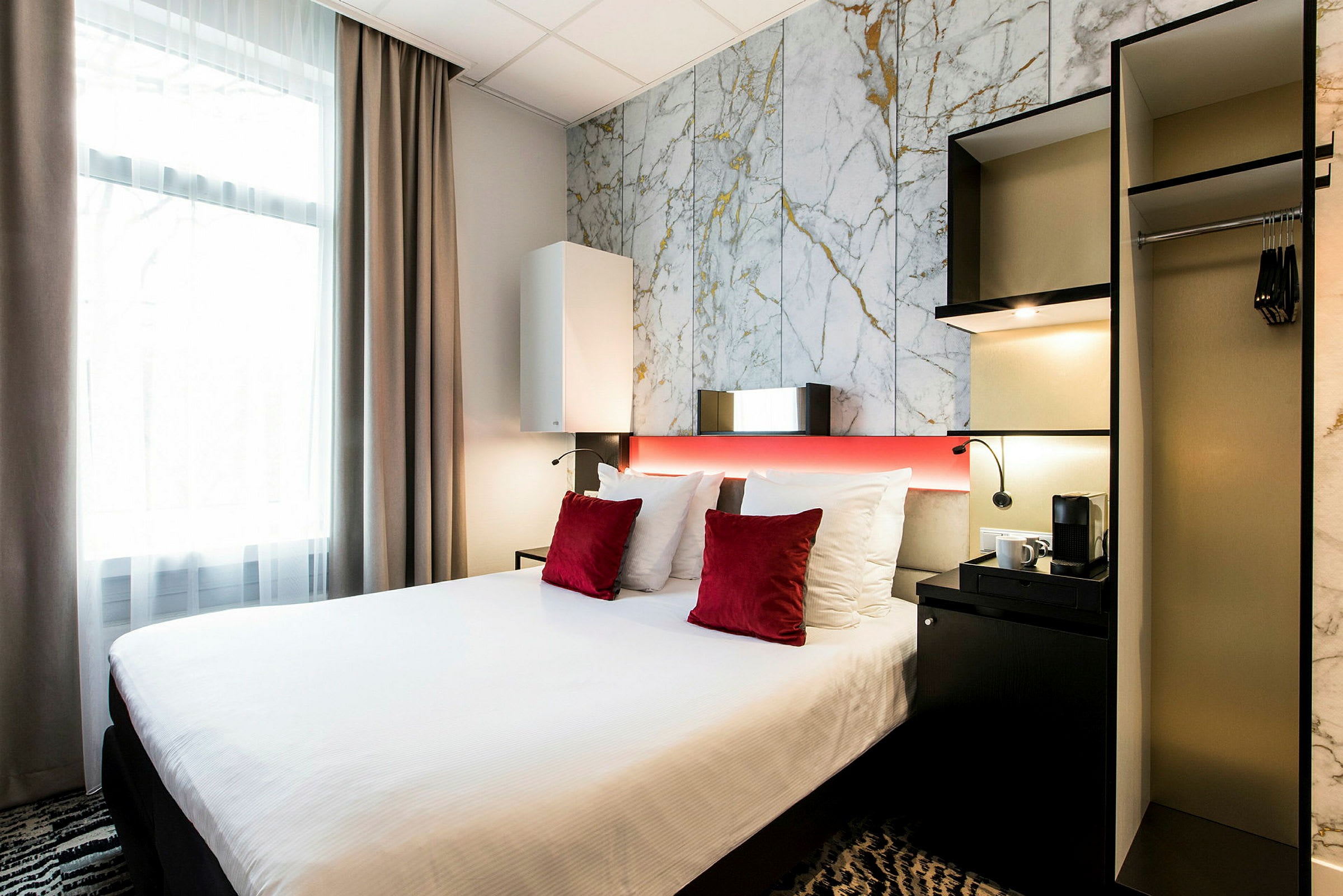 Et hotellrom med en seng med hvitt sengetøy og røde pynteputer, ved siden av et skap og et lite bord med en kaffemaskin. (AI-generert tekst)
