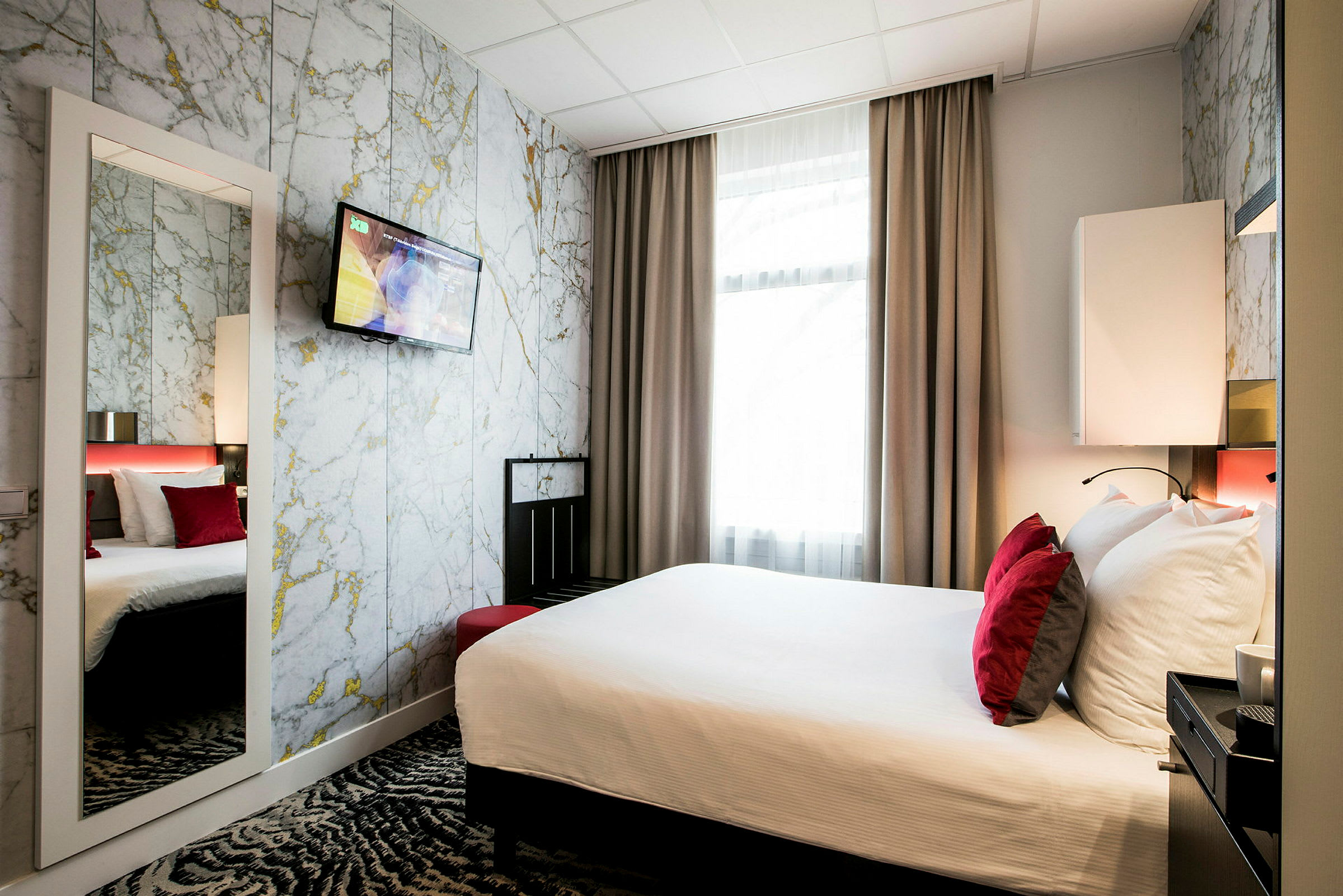 Et hotellrom med en dobbeltseng med hvitt sengetøy og røde puter foran et marmorert veggmønster og en veggmontert TV. (AI-generert tekst)