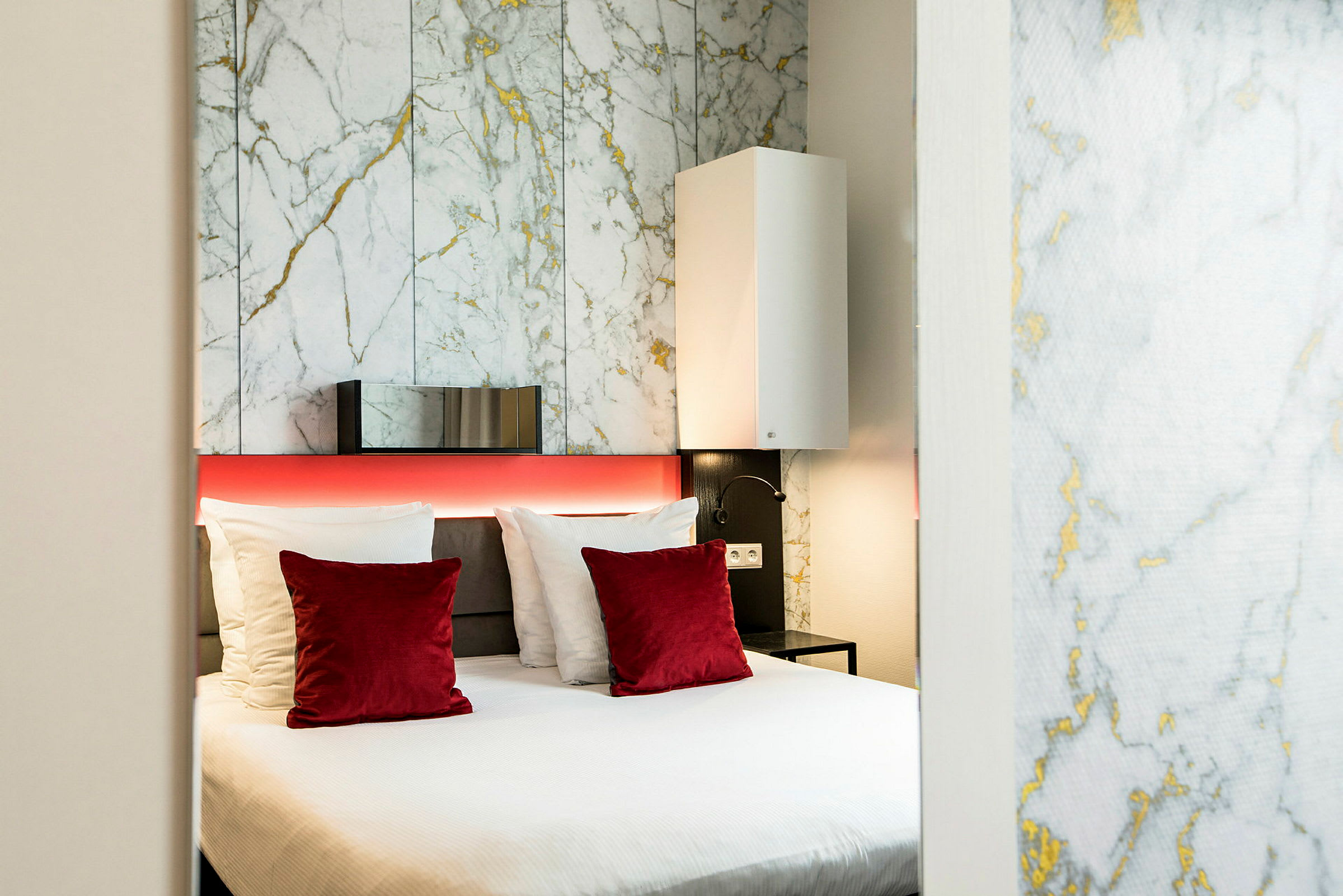 Et moderne hotellrom med en dobbeltseng, hvite og røde puter samt en bakgrunnsvegg med marmormønster. (AI-generert tekst)