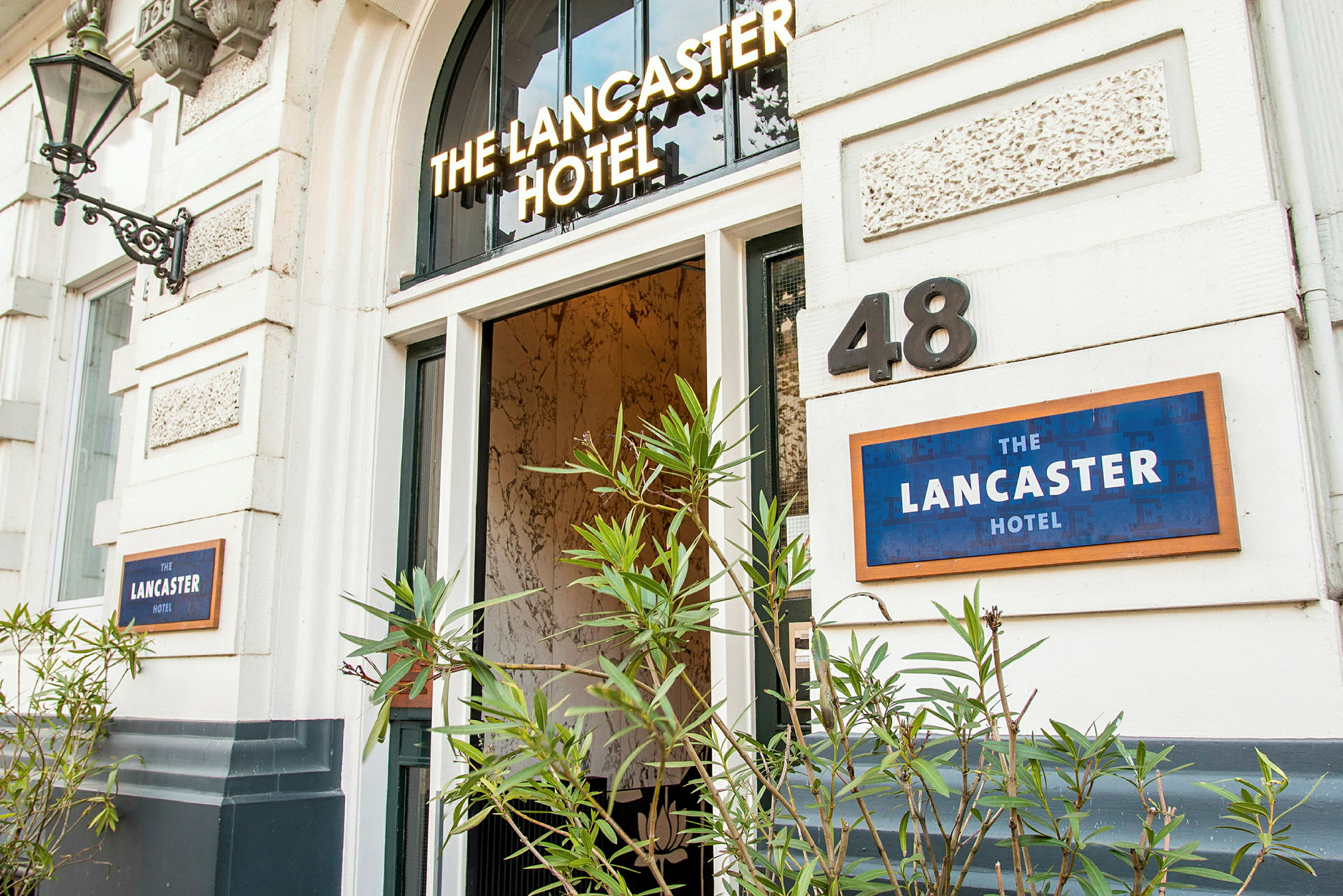 Inngang til et hotell med navnet 'The Lancaster Hotel', omkranset av grønne planter. (AI-generert tekst)
