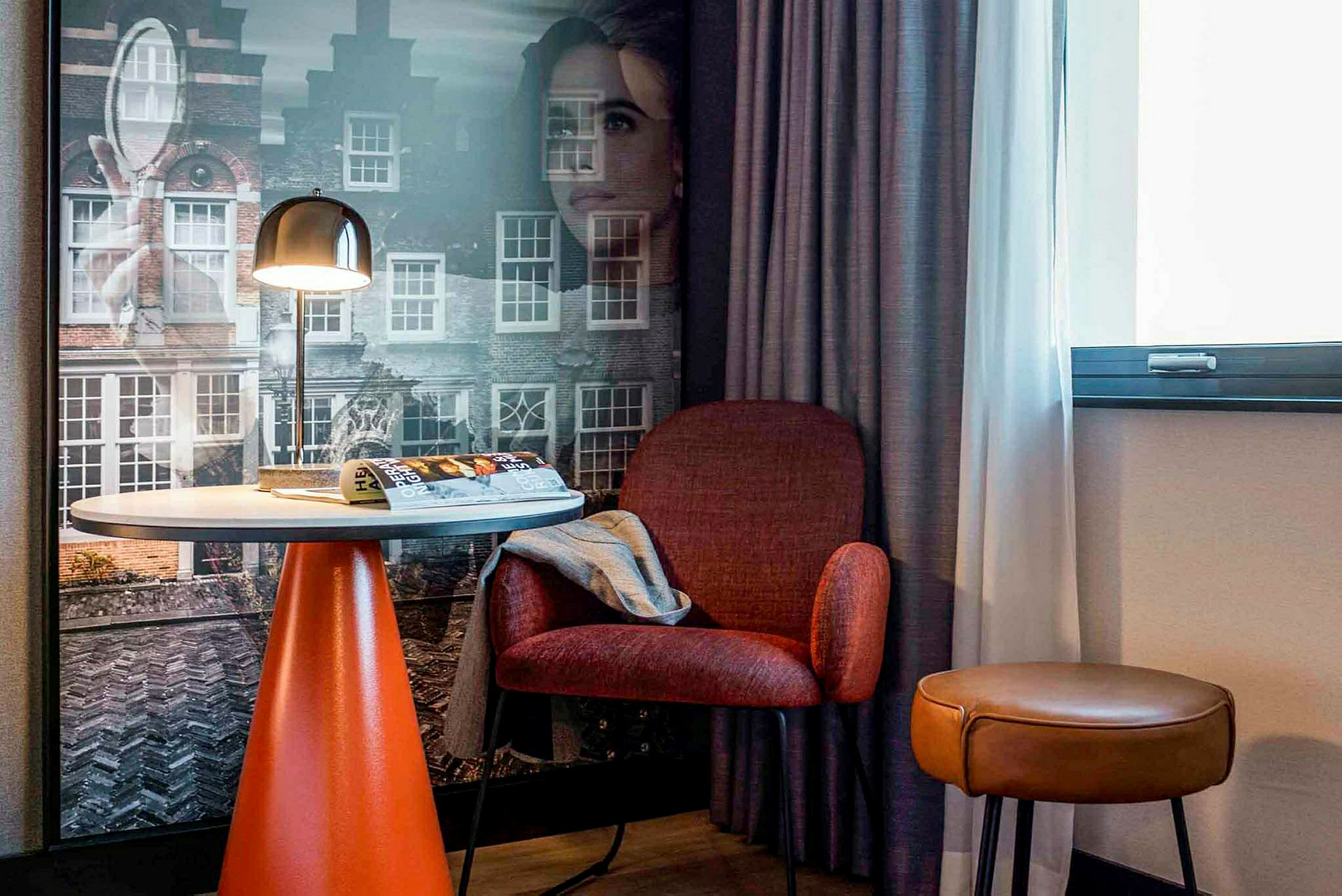 Et hotellrom med et bord, en rød stol og en brun krakk foran et vindu og en vegg med bymotiv. (AI-generert tekst)
