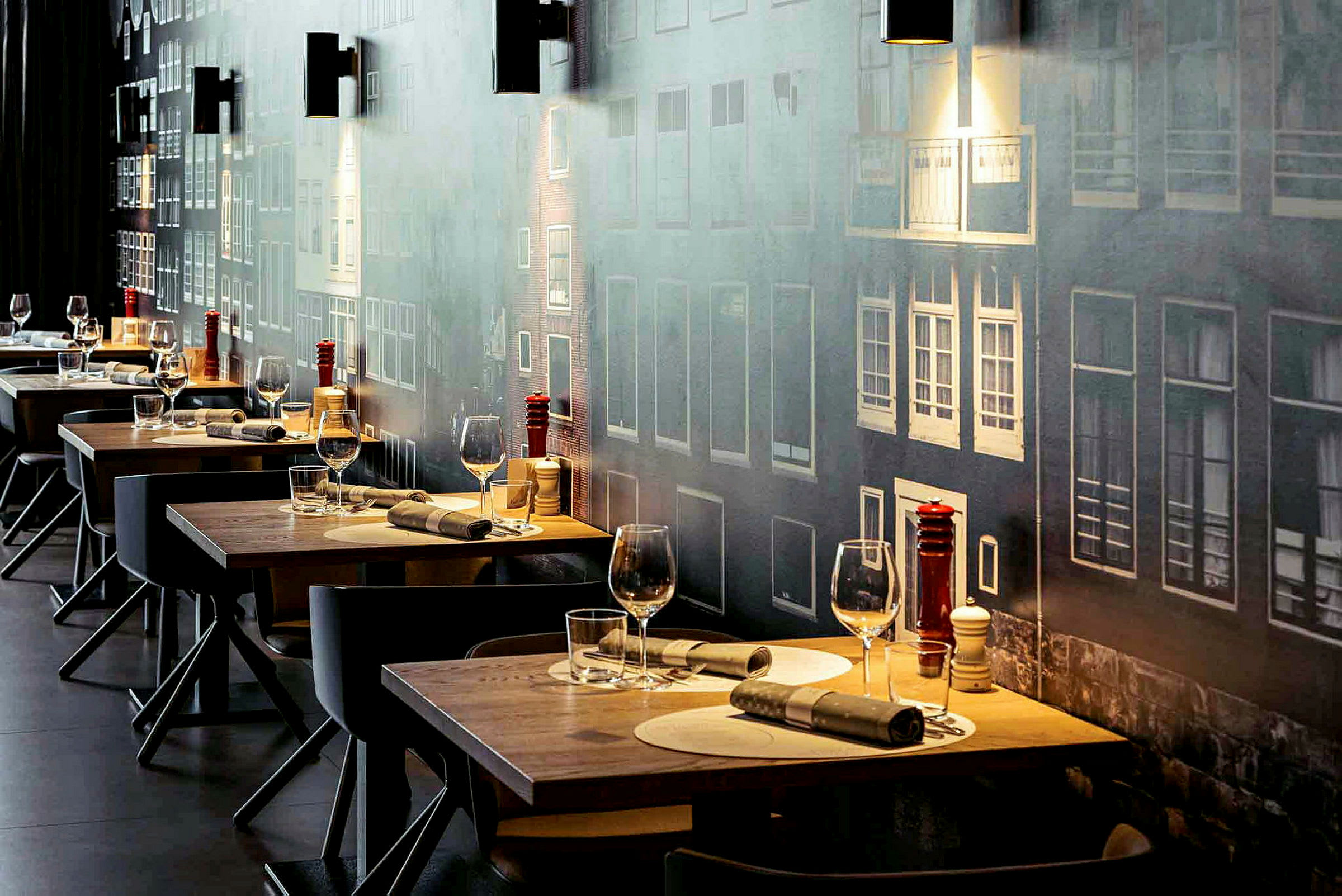 Flere dekkede restaurantbord med glass og bestikk, mot en vegg med fasademotiv. (AI-generert tekst)