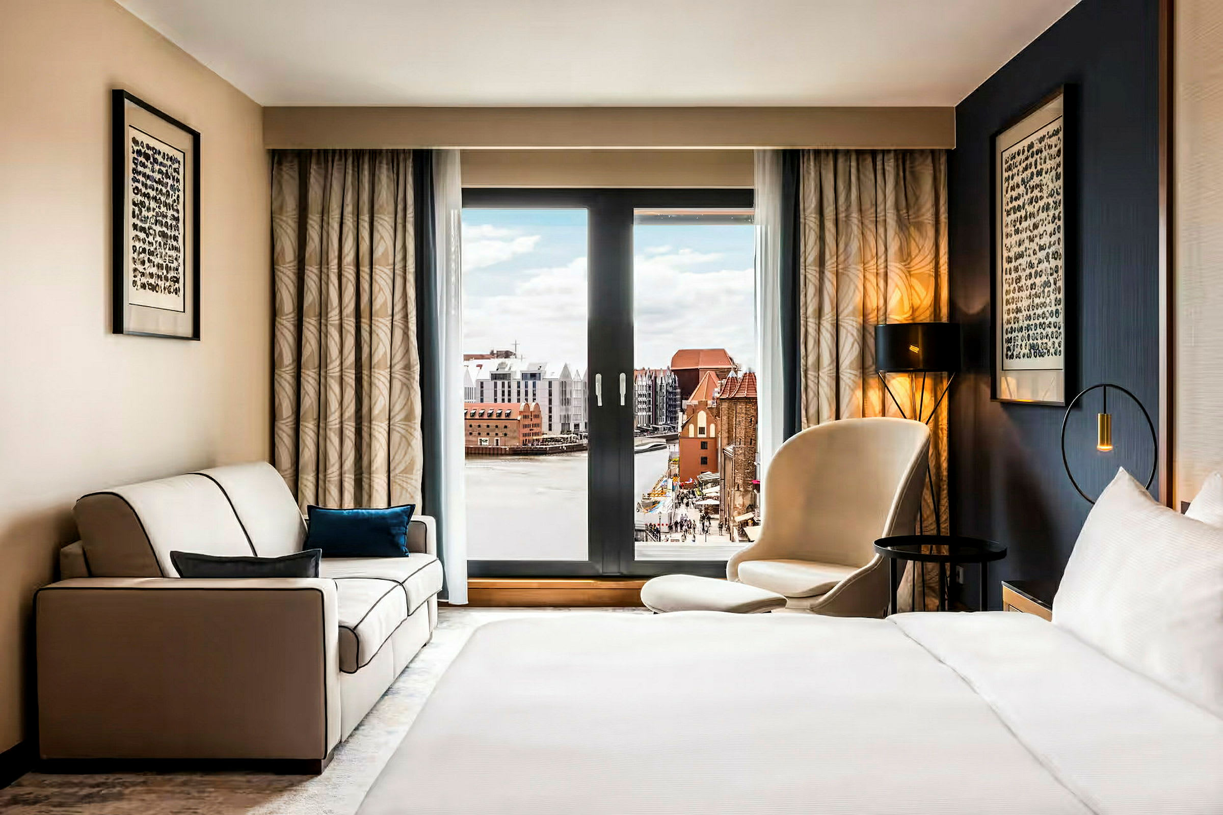 Et hotellrom med en seng, en sofa og en stol foran et stort vindu med utsikt over en by og vann. (AI-generert tekst)
