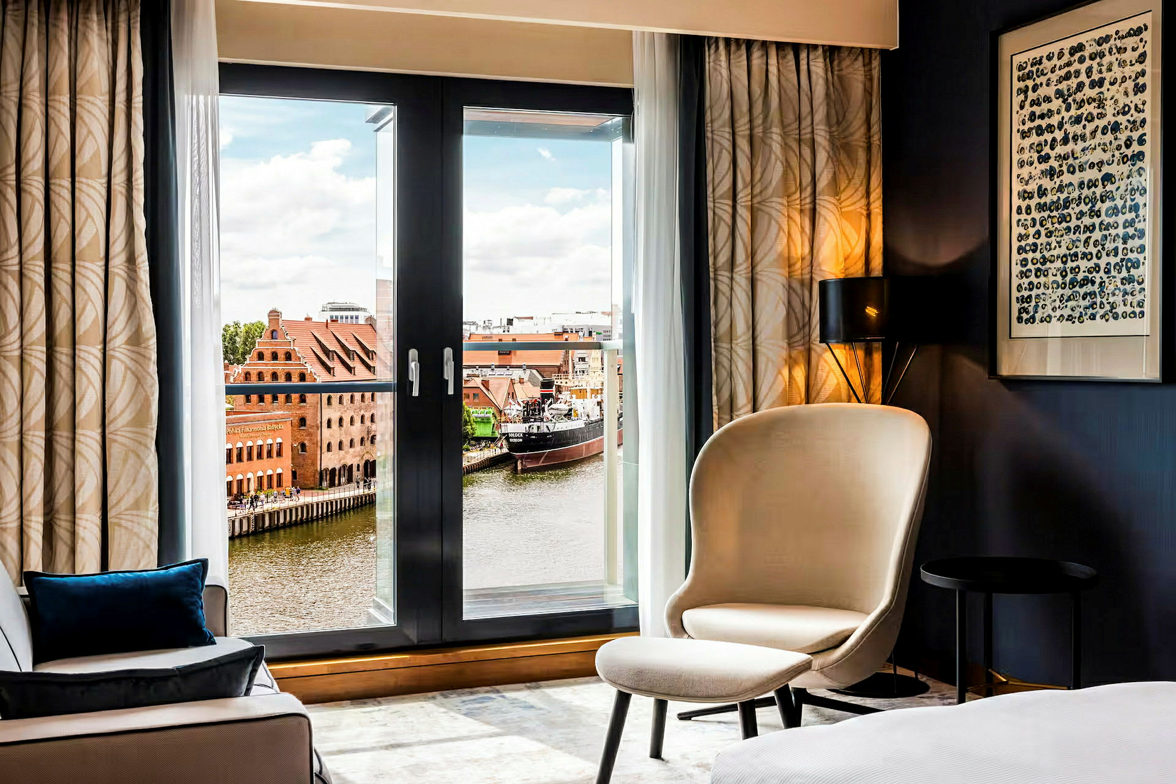 Et hotellrom med en lenestol og utsikt over en elv med bygninger og en båt utenfor. (AI-generert tekst)