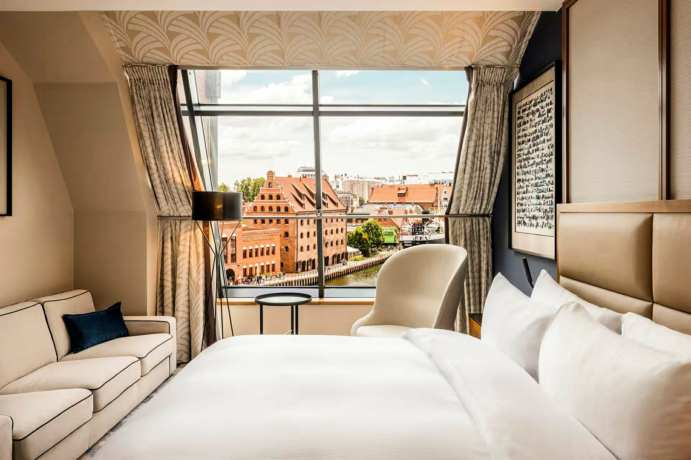 Et hotellrom med en seng, sofa og lenestol, samt et stort vindu med utsikt over en bymiljø med røde teglbygninger. (AI-generert tekst)