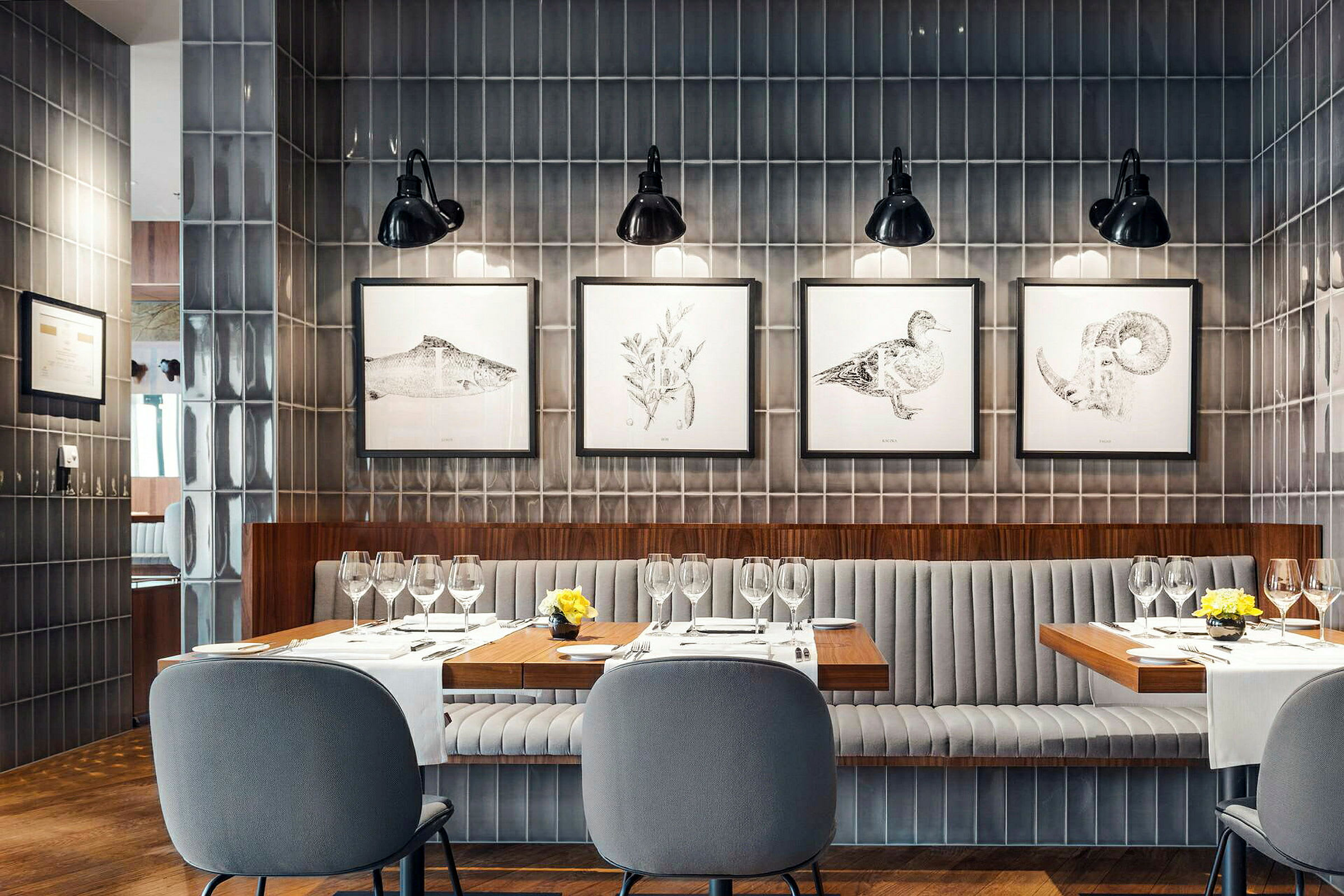En moderne restaurant med grå veggpaneler, et bord dekket med glass og servietter, samt stoler med polstrede ryggstøtter. Malerier henger på veggen bak. (AI-generert tekst)