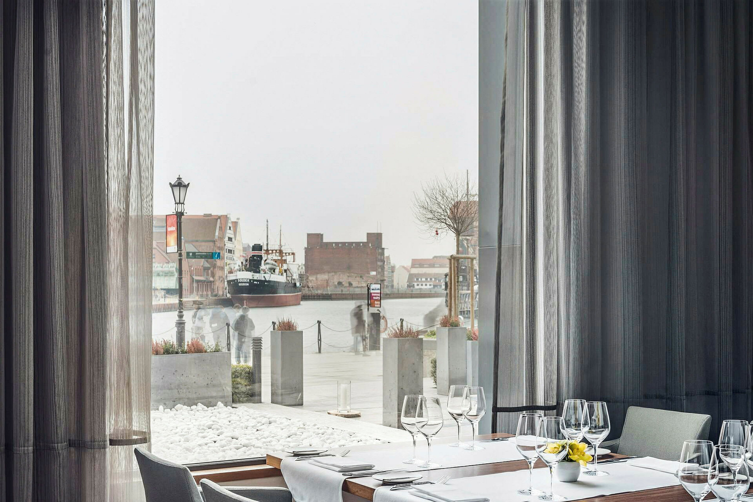 Restaurantbord ved et stort vindu med utsikt over en havn med et skip. (AI-generert tekst)