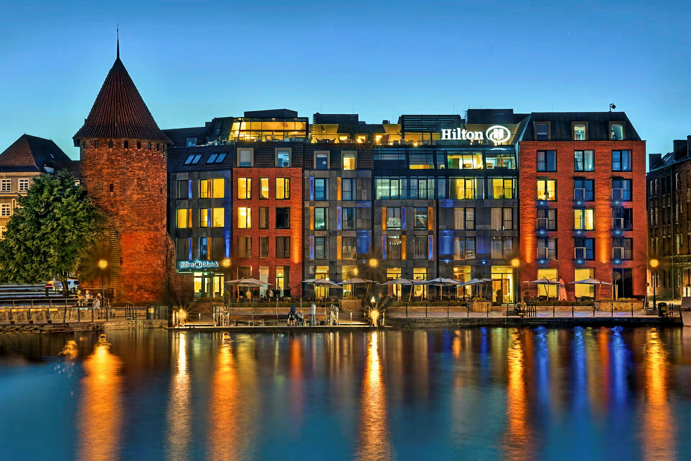 En historisk murbygning ved en moderne hotell ved vannet om kvelden. (AI-generert tekst)