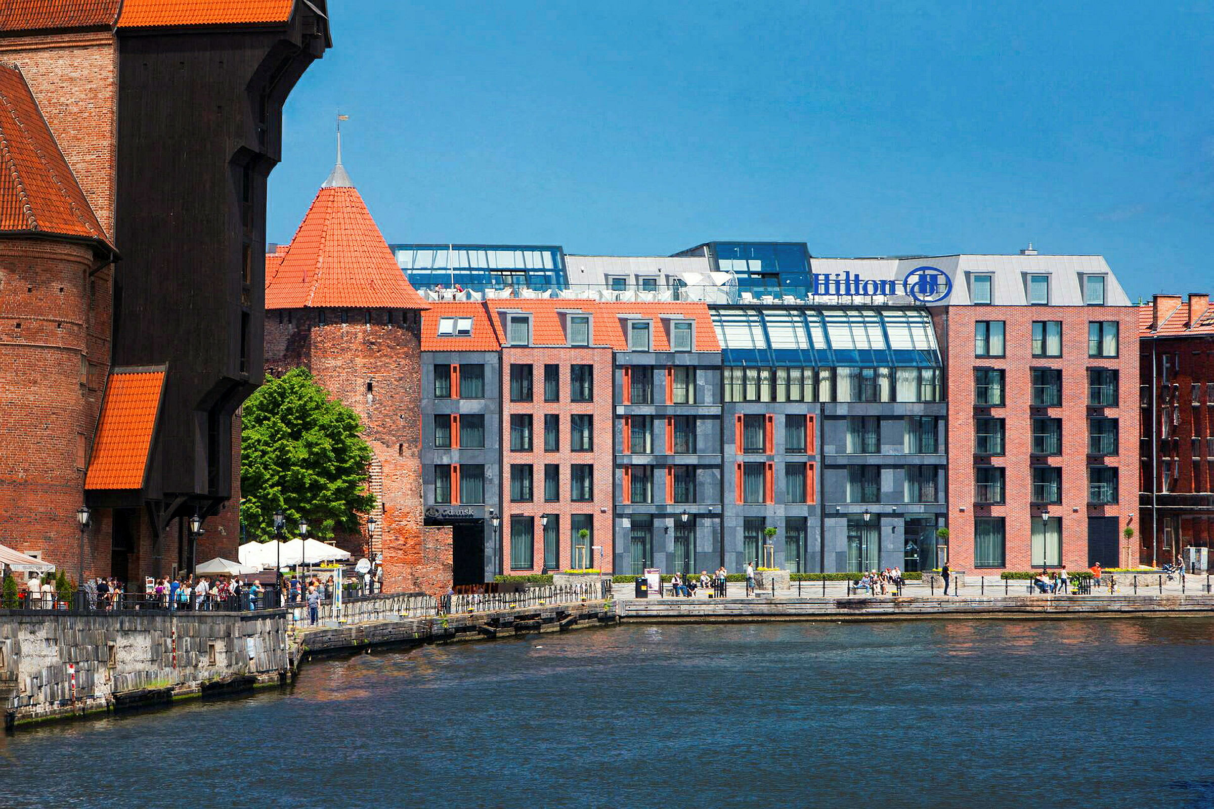 Et vannløp med bygninger i bakgrunnen, inkludert et hotell med moderne glassfasade og historiske teglkonstruksjoner. (AI-generert tekst)