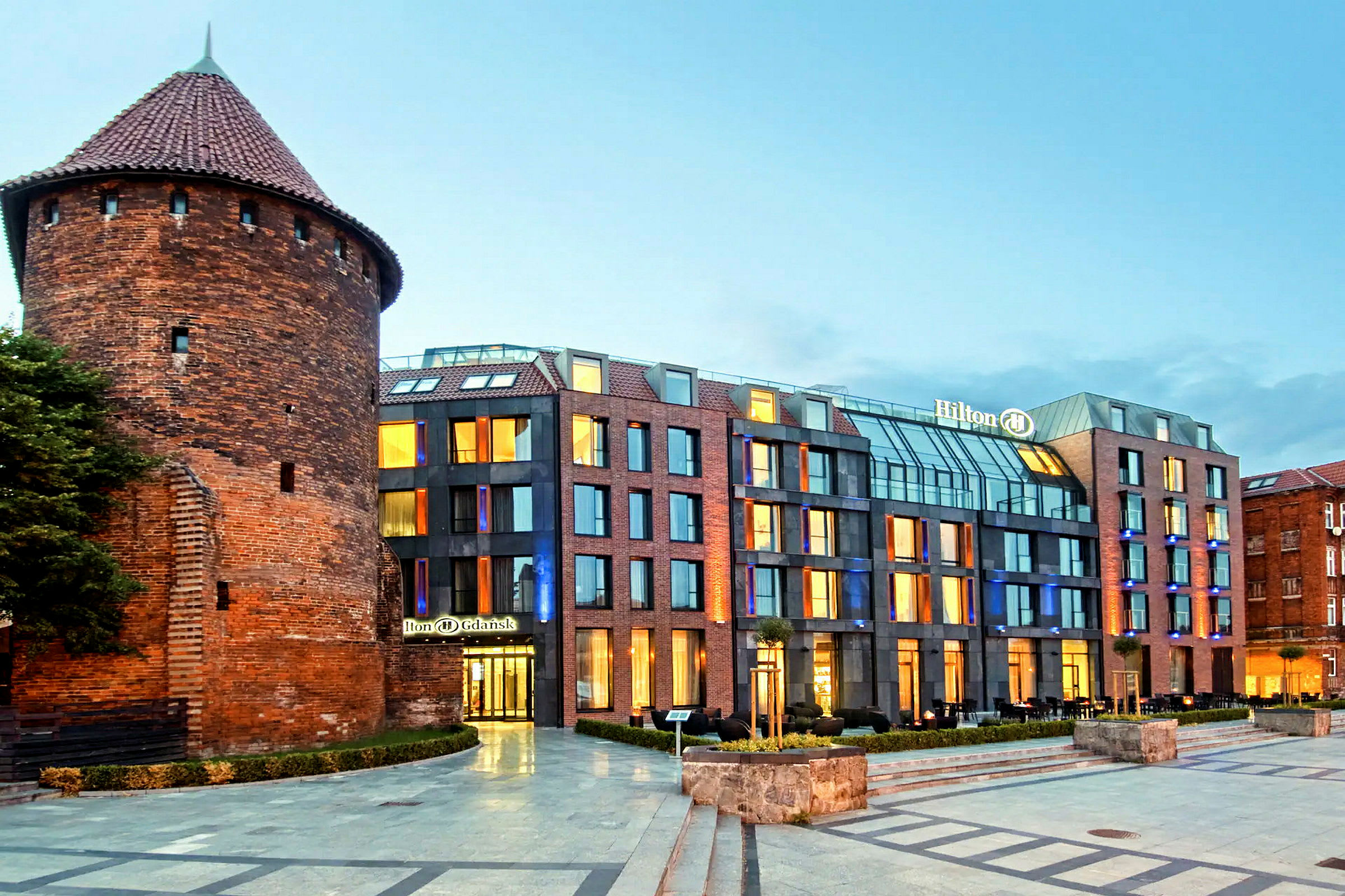 Et moderne hotell ved siden av en gammel rund mursteinsbygning i bymiljø. (AI-generert tekst)