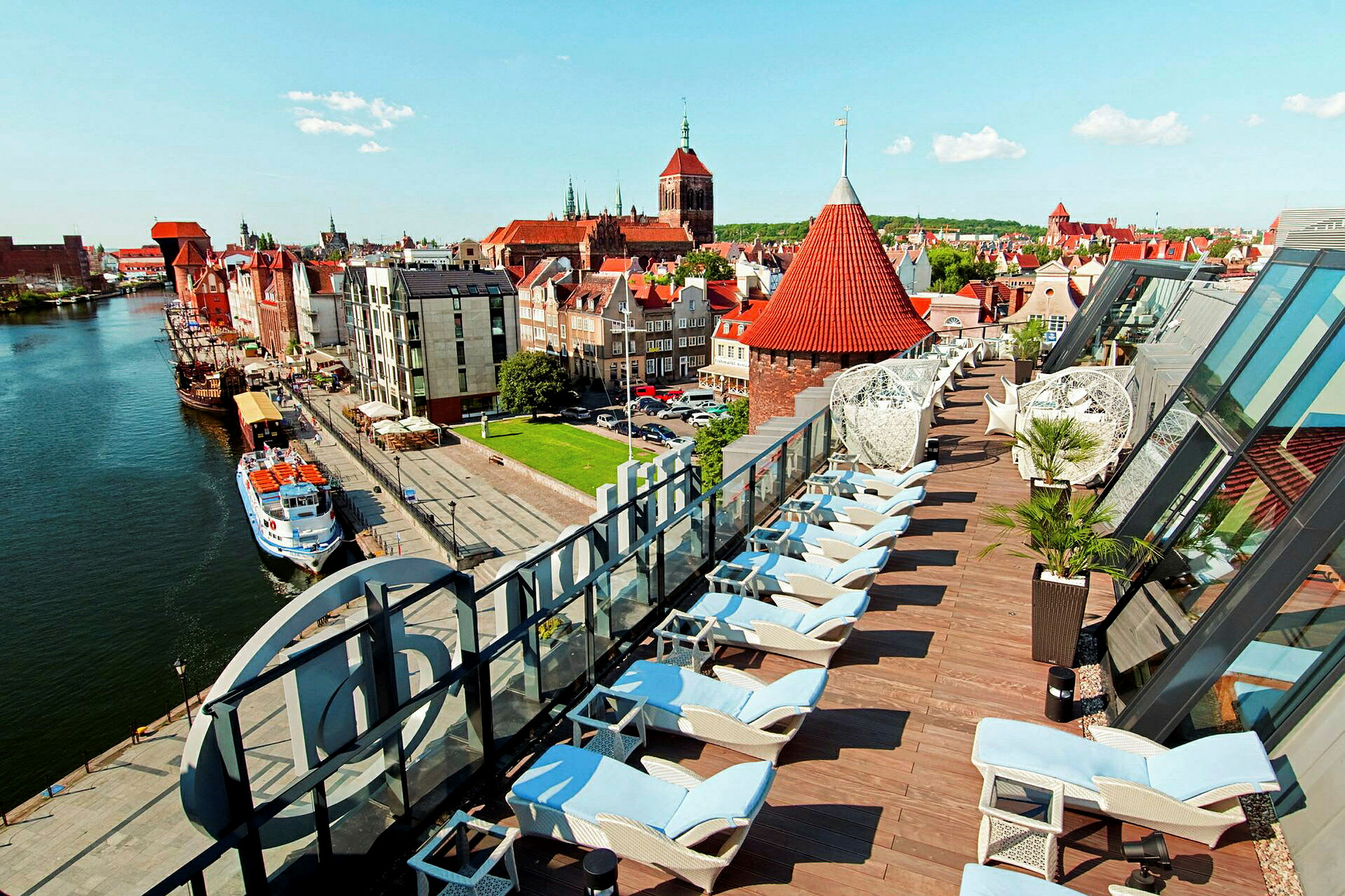 Bilde av en by med elv, bygninger og en takterrasse med solsenger. (AI-generert tekst)