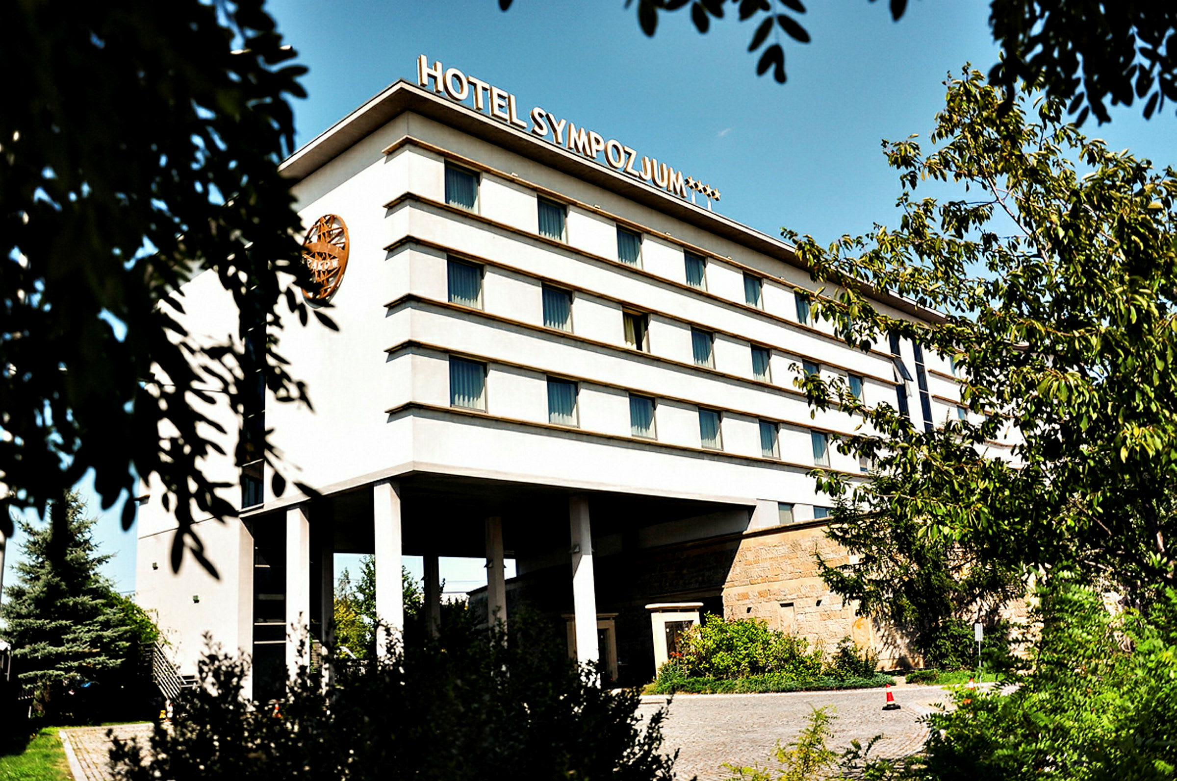 Bygning med flere etasjer, merket med teksten 'Hotel Sympozjum', omgitt av trær og vegetasjon. (AI-generert tekst)