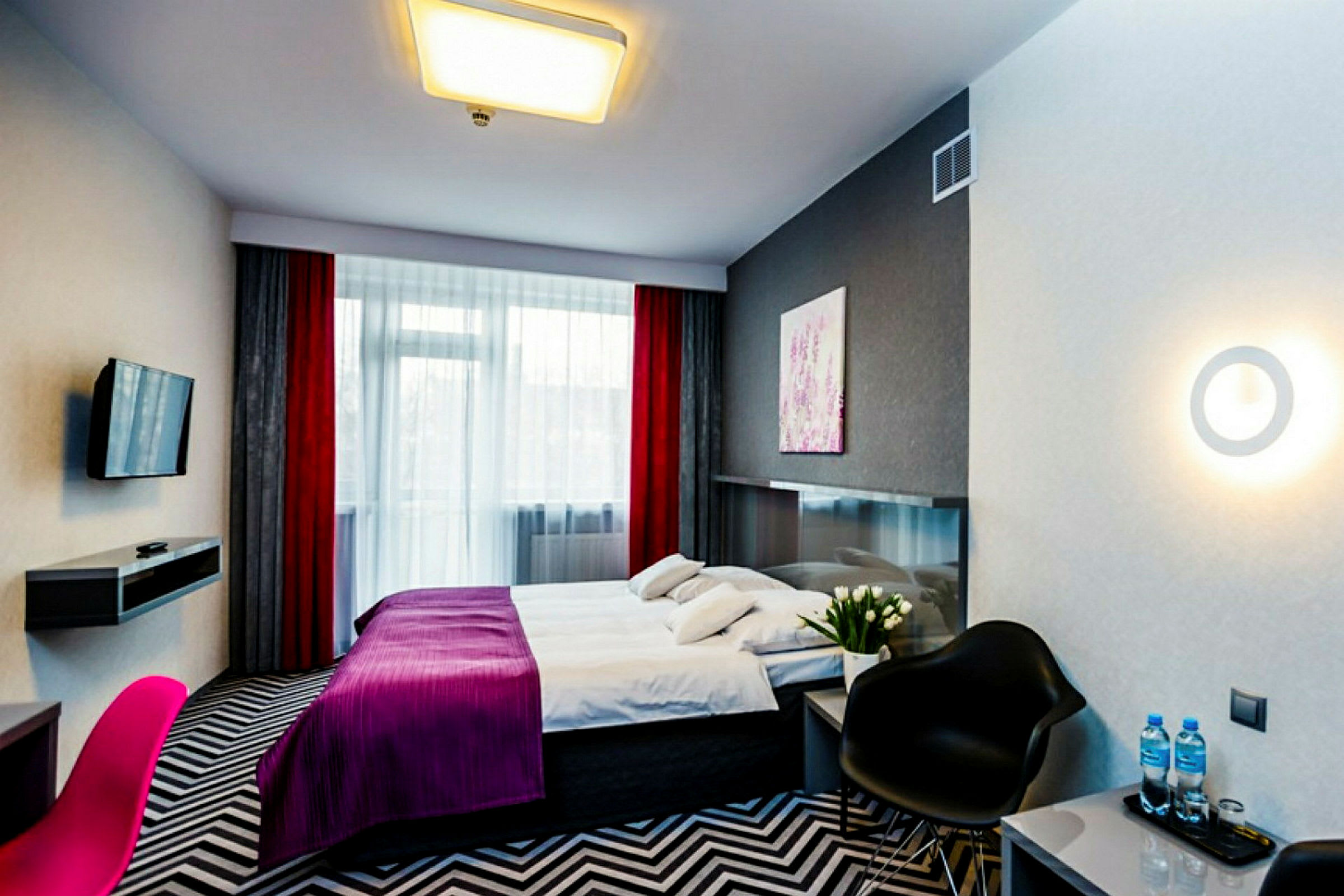 Et moderne hotellrom med en stor seng, fargerike gardiner og en TV montert på veggen. Rommet har også en stol, et bord og dekorativ belysning. (AI-generert tekst)
