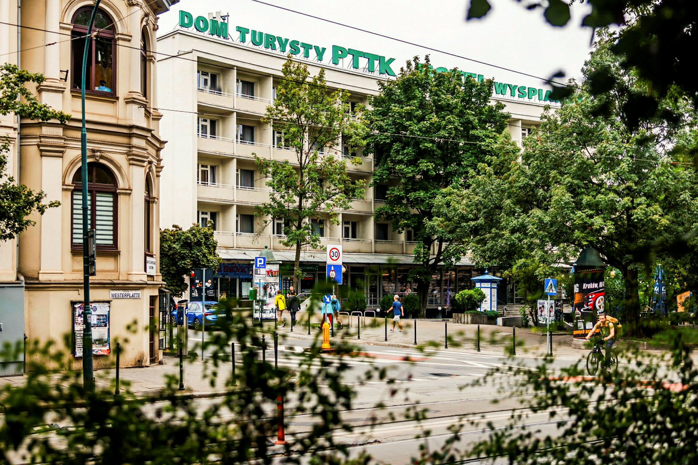 Et bilde av en bygning med skilt "Dom Turysty PTTK Hotel Wyspiański" omgitt av trær. På gaten foran sees mennesker, sykler og skilt. (AI-generert tekst)