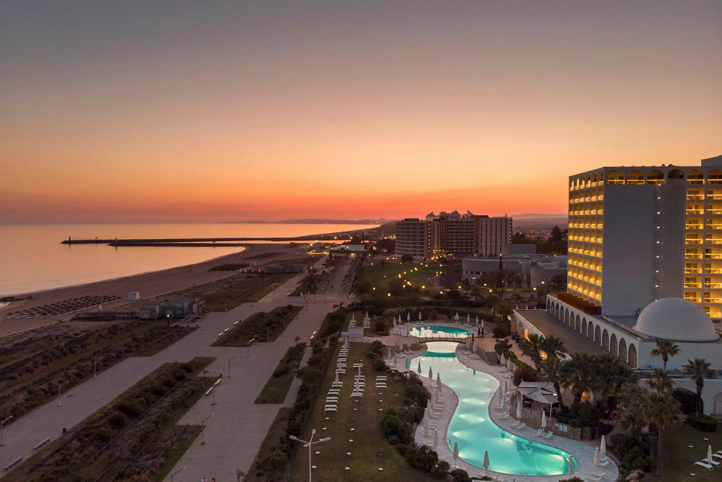 En utsikt over en strandpromenade, hotell med opplyste vinduer og basseng ved solnedgang. (AI-generert tekst)