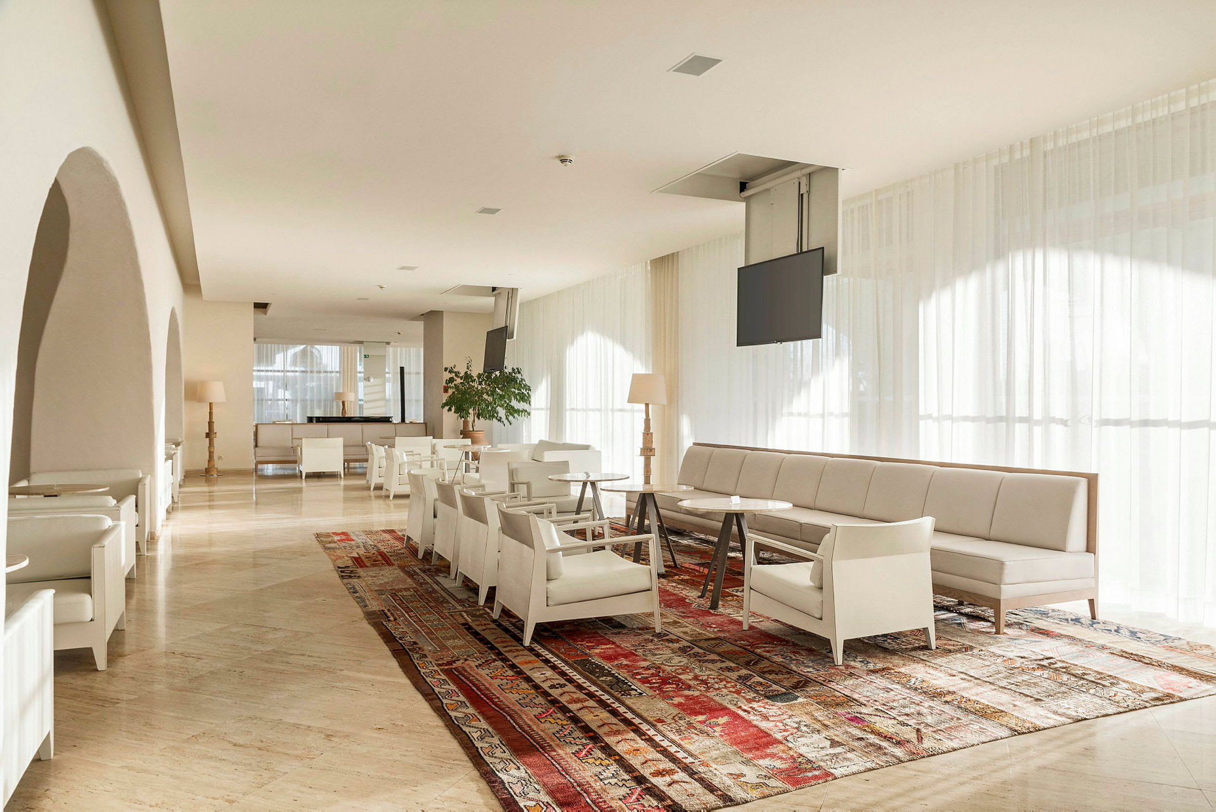 Et lyst og moderne loungeområde med sofaer, lenestoler og et langt bord på et fargerikt teppe. Store vinduer med lyse gardiner slipper inn naturlig lys. (AI-generert tekst)