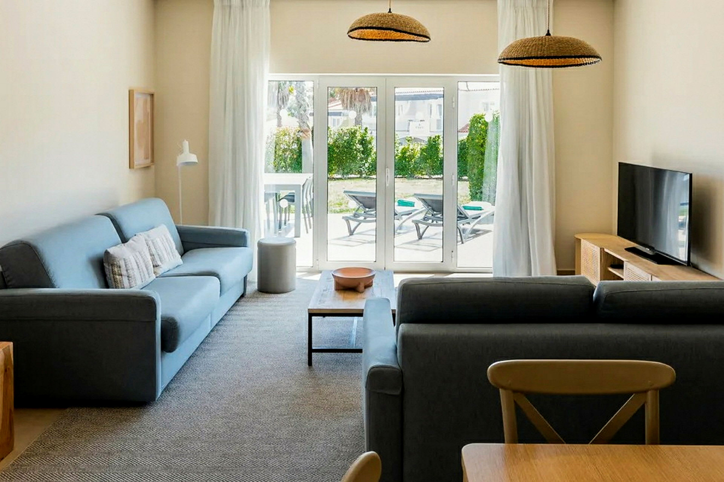 En stue med to sofaer, et salongbord, en TV og en glassdør som vender ut mot en terrasse med solsenger. (AI-generert tekst)