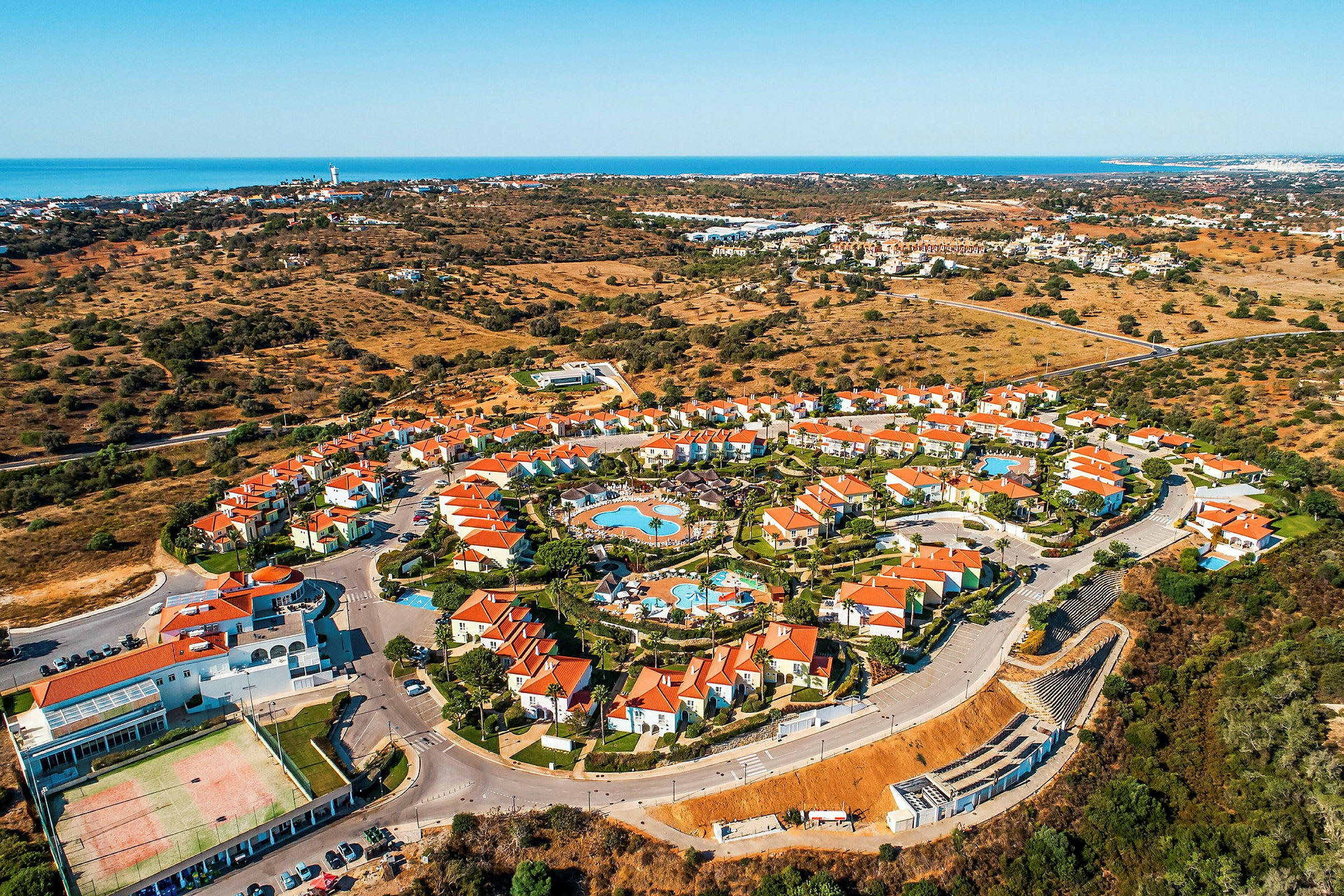 Luftfoto av et feriested med røde tak, omgitt av landskap og havet i bakgrunnen. (AI-generert tekst)