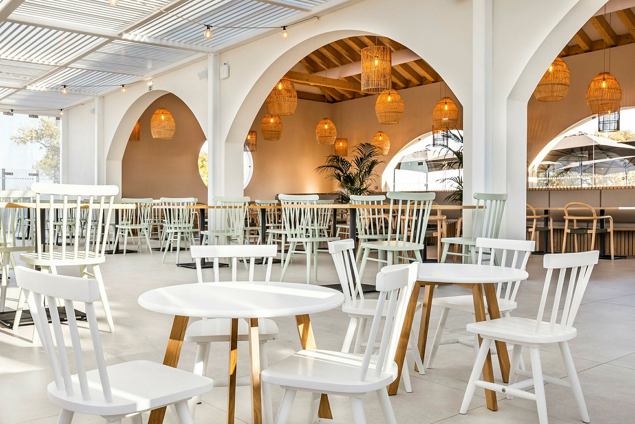 En lys restaurant med hvite trebord og stoler samt hengende kurvlamper. (AI-generert tekst)