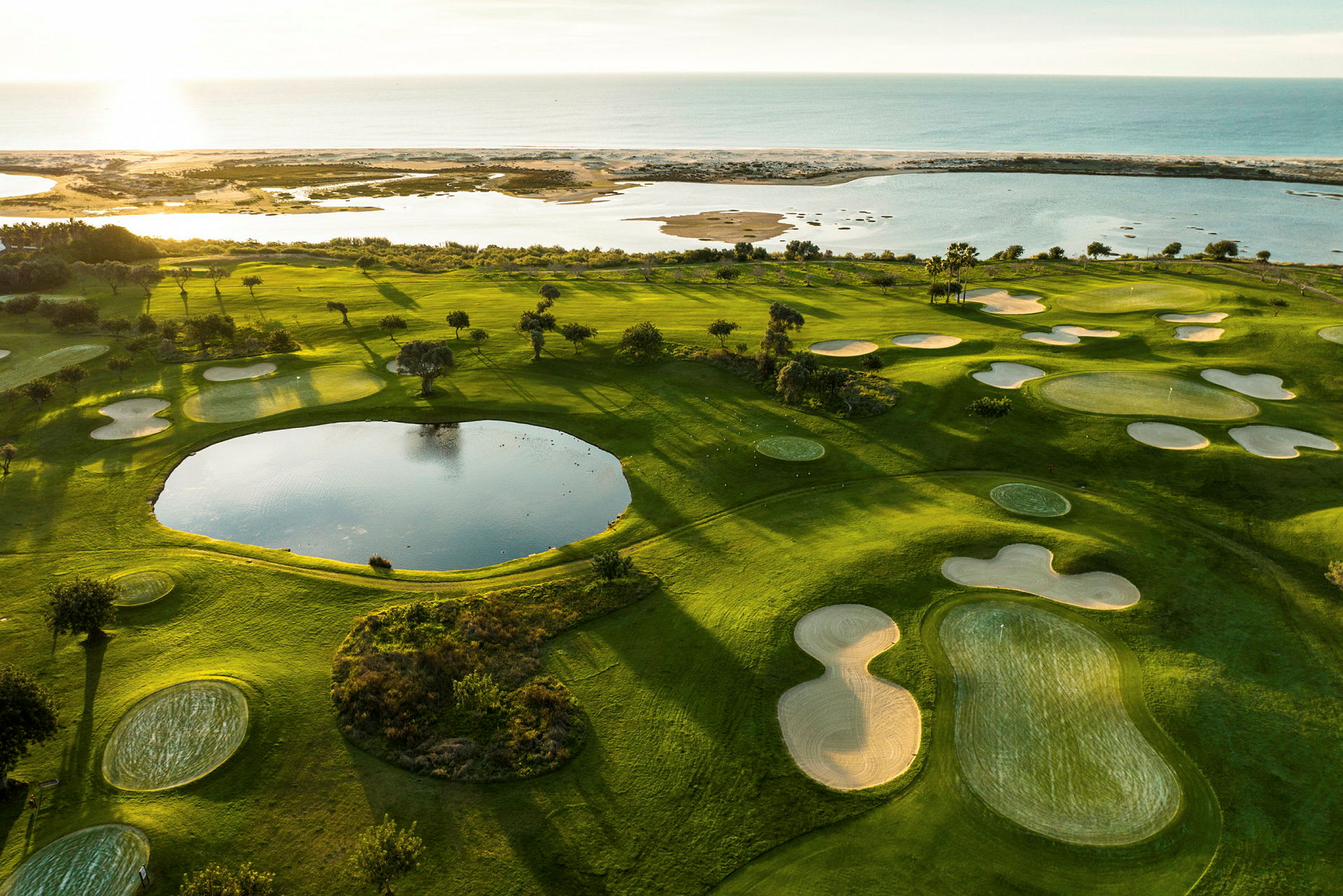 Utsikt over en grønn golfbane med sandbunkere og en dam, nær kysten og havet. (AI-generert tekst)